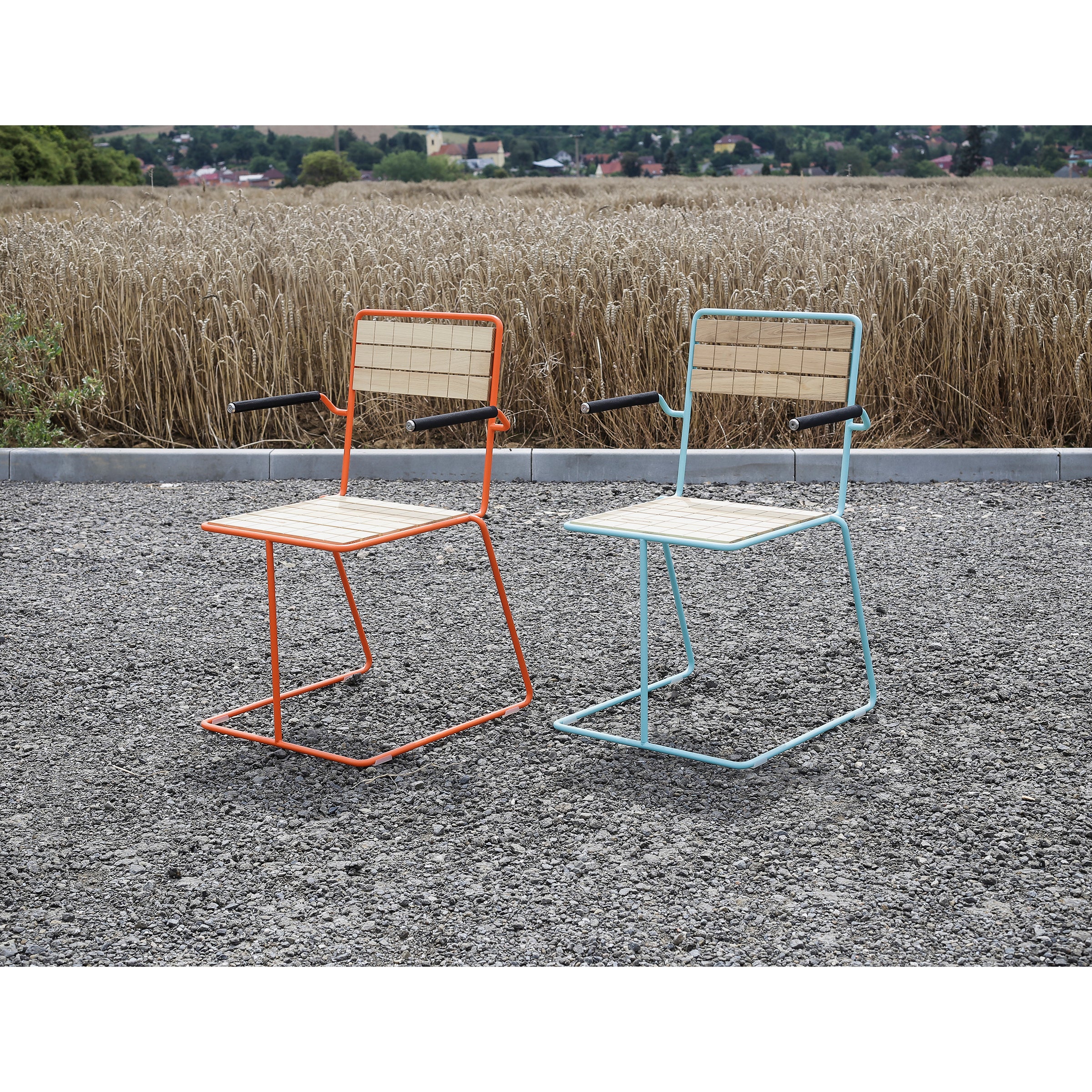 Two modern Tina Armchairs by Egoe, with wire frames and wooden seats—one orange, the other light blue—rest on a gravel path. This stylish outdoor furniture is perfectly positioned before a wheat field with trees and buildings enhancing the backdrop.