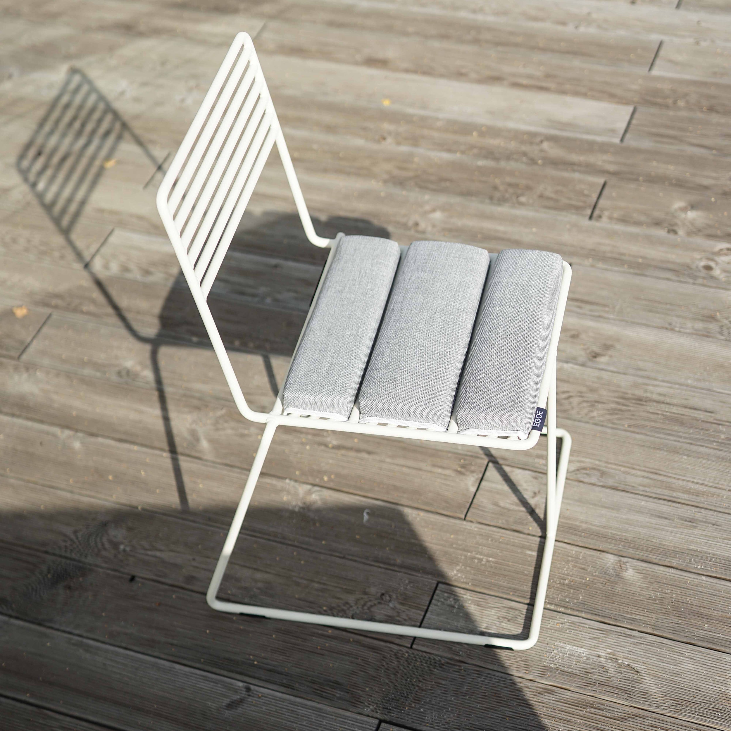 A white Tina Chair by Egoe, with a minimalist design and powder-coated finish, adorns the wooden deck. It features a slatted backrest and light gray cushioned seat, elegantly casting its shadow in the sunlight.