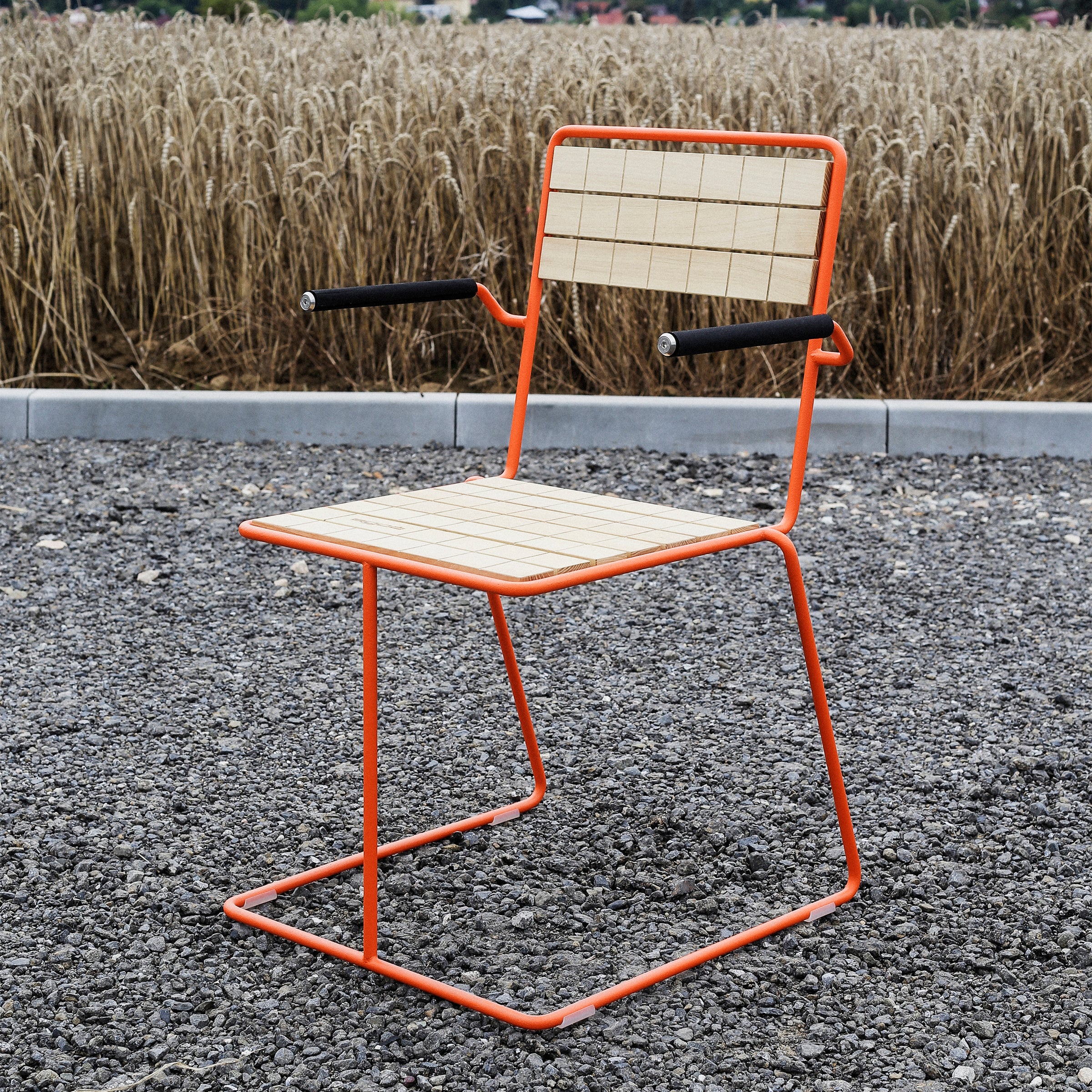 The Tina Armchair by Egoe features a contemporary design with an orange powder-coated metal frame, wooden seat and backrest, and armrests. Ideal for outdoor settings, it’s beautifully displayed on gravel against a wheat field backdrop.