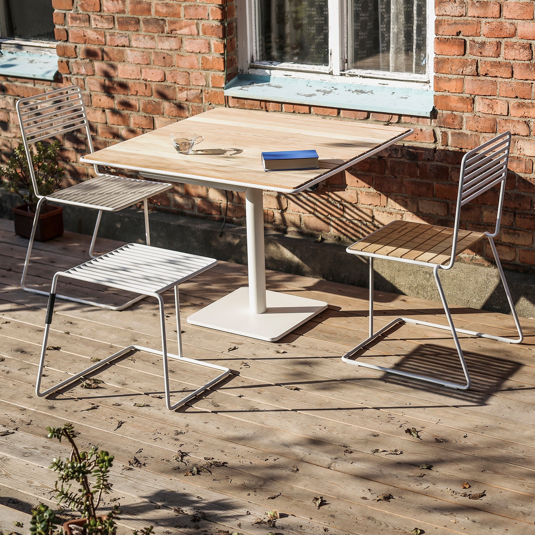 A wooden patio featuring the Egoe Tina Table and two metal chairs creates a visually light aura. The table is adorned with a blue book and glass ashtray, while sunlight casts shadows on the floor. A brick wall with a window completes this serene scene.