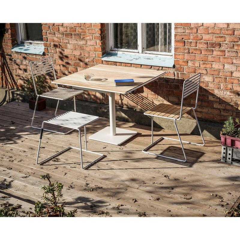 A charming wooden patio features a square Tina Series table with three metal Tina Chairs from Egoe. A blue book and glass cup are on the sturdy, powder-coated table. The space is elegantly bordered by a brick wall, accented with potted plants on the wooden floor.