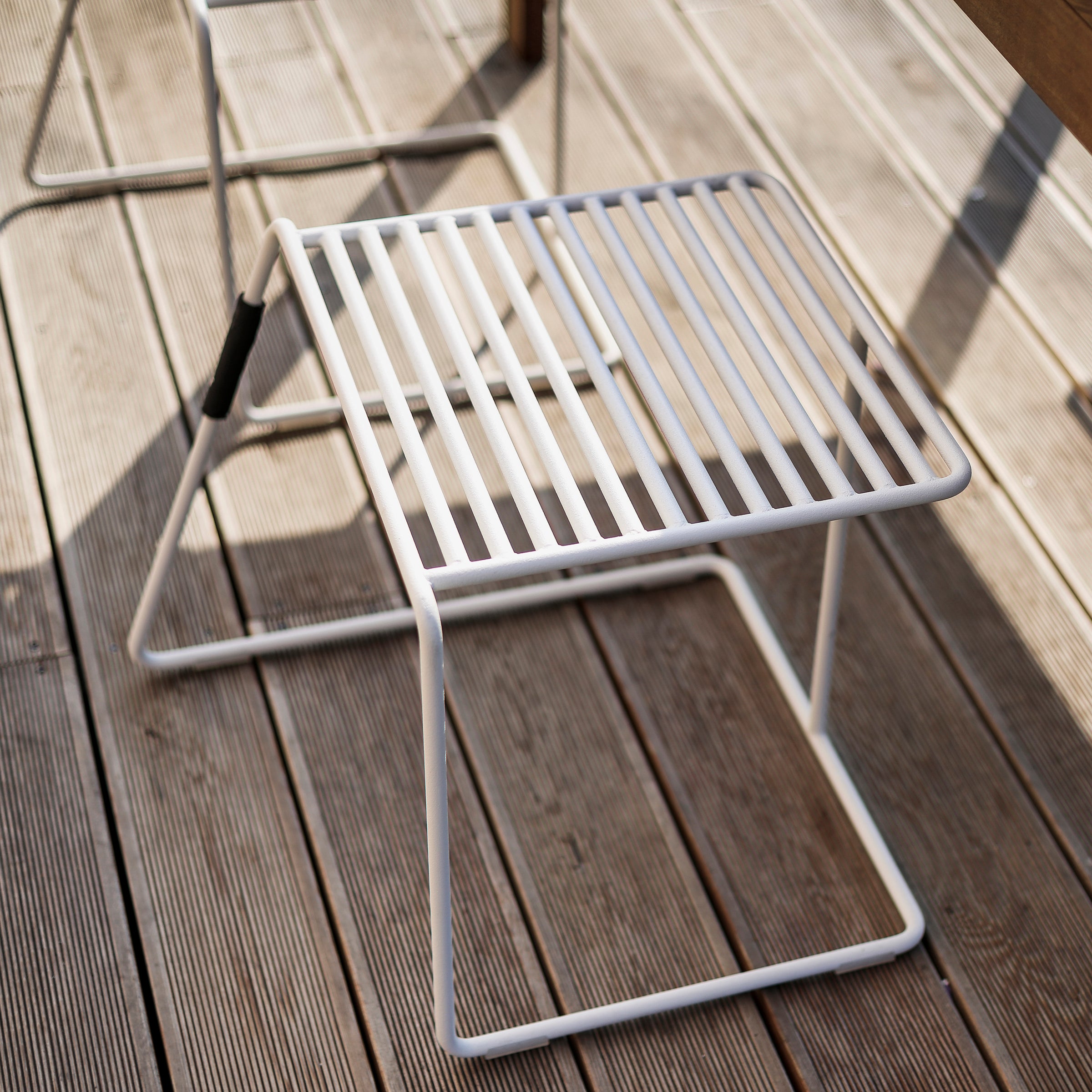 The Egoe Tina Stool, a minimalist white metal piece with a slatted seat and three legs, rests on a wooden deck. Shadows imply sunlight is streaming through, making it ideal for outdoor seating.