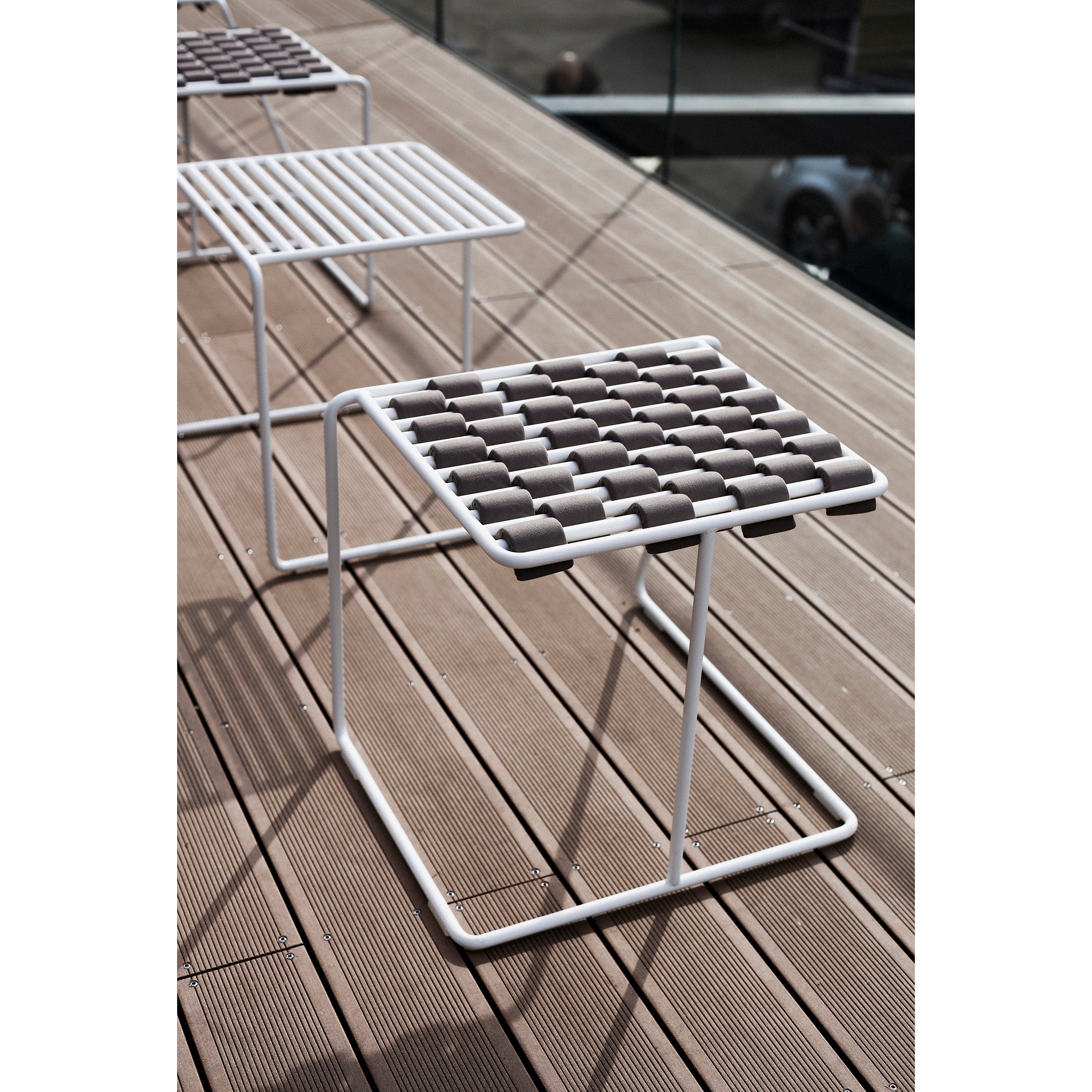 Three modern Tina Stools from Egoe, with grid-pattern seats and a sleek three-legged design, are on a wooden deck. Chair shadows cast striped patterns on the surface, while glass railings elegantly frame the seating area.