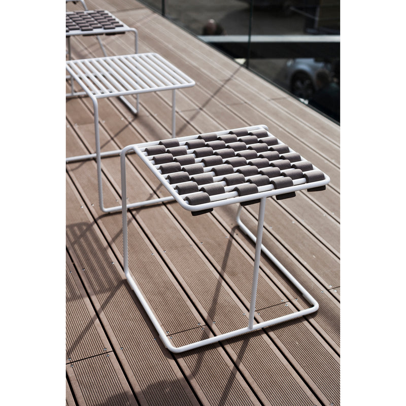 Three modern Tina Stools from Egoe, with grid-pattern seats and a sleek three-legged design, are on a wooden deck. Chair shadows cast striped patterns on the surface, while glass railings elegantly frame the seating area.