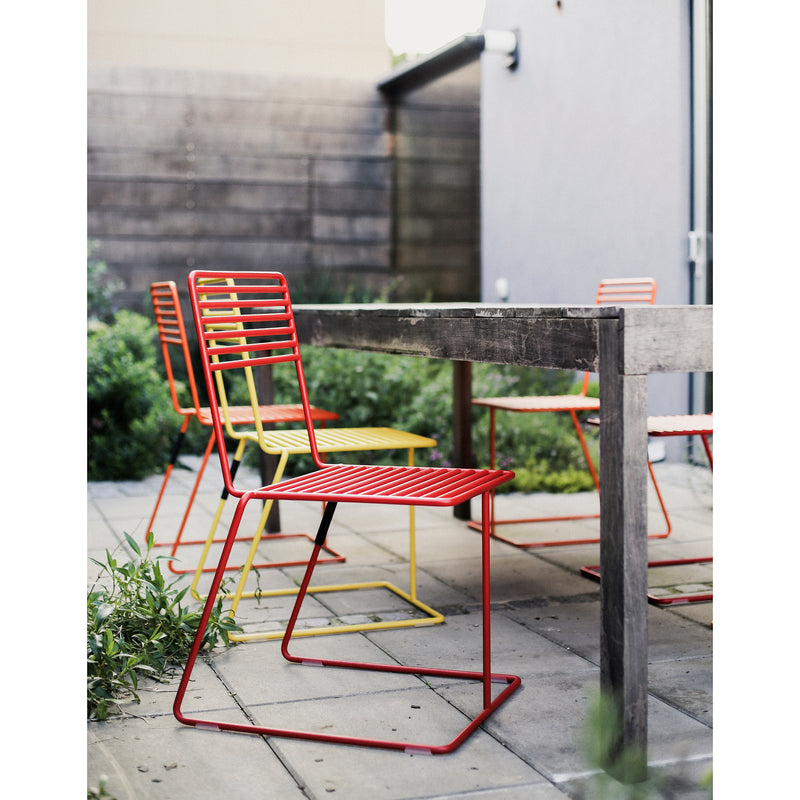 Four Tina Chairs by Egoe, two in red and two in yellow with a powder-coated finish, surround a rectangular wooden table on a patio. The modern setting is complemented by greenery and wooden fencing for privacy.