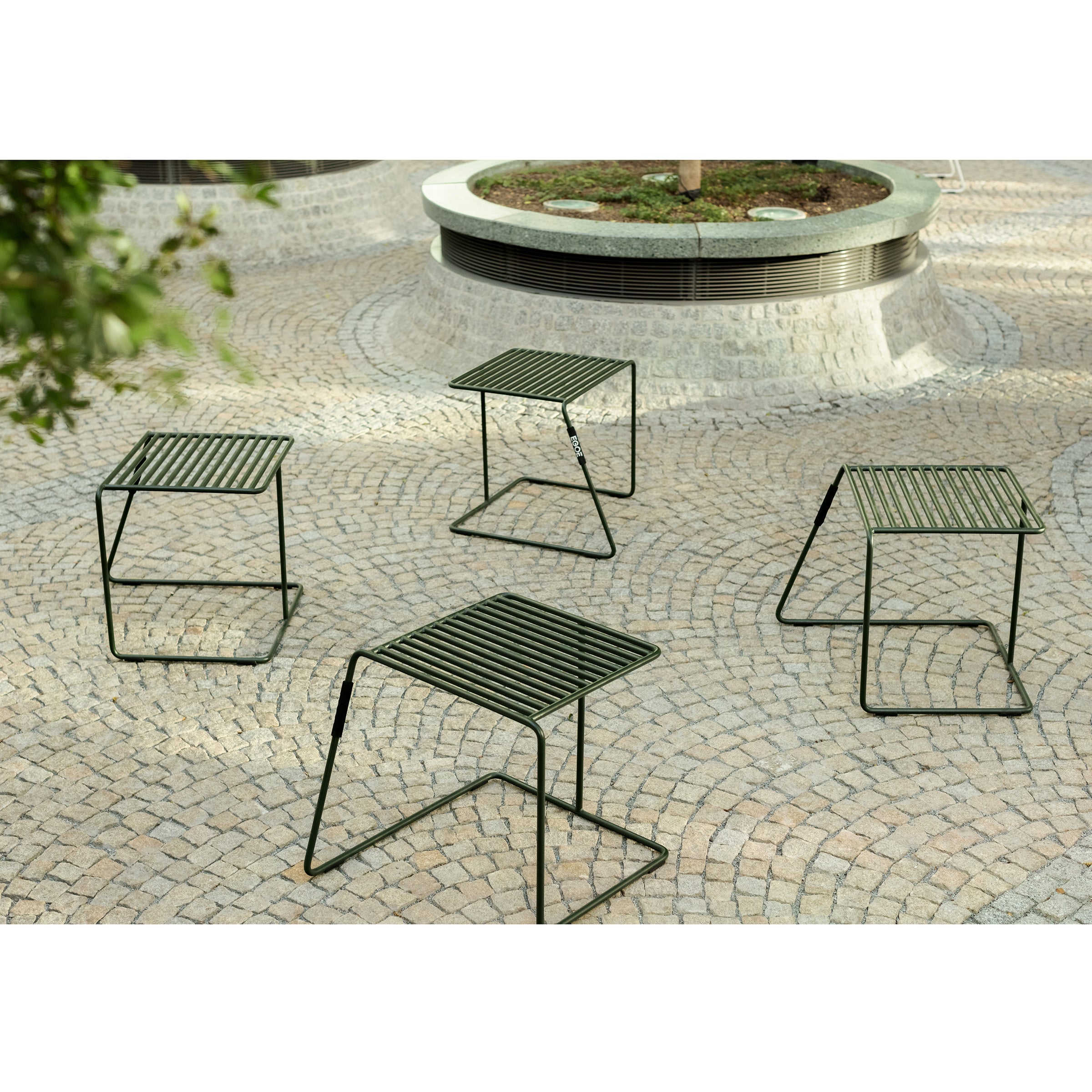 Four green metal Tina stools by Egoe with slatted tops are set on a cobblestone patio. A raised circular stone planter with greenery is partially visible in the background, enhancing the peaceful outdoor ambiance.