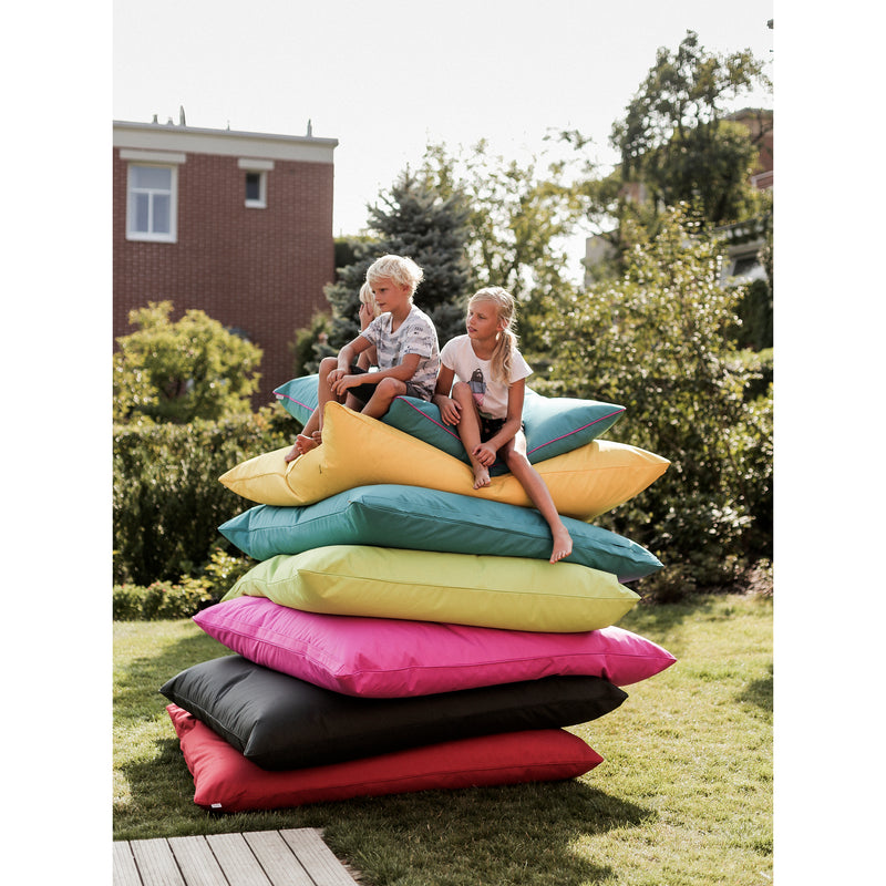 Two children sit atop a tall stack of colorful Vago Bean Bag cushions by Egoe, resembling playful seats, in a sunny garden. The cushions display shades of red, pink, yellow, green, and black. Trees and a brick building frame this delightful outdoor scene.