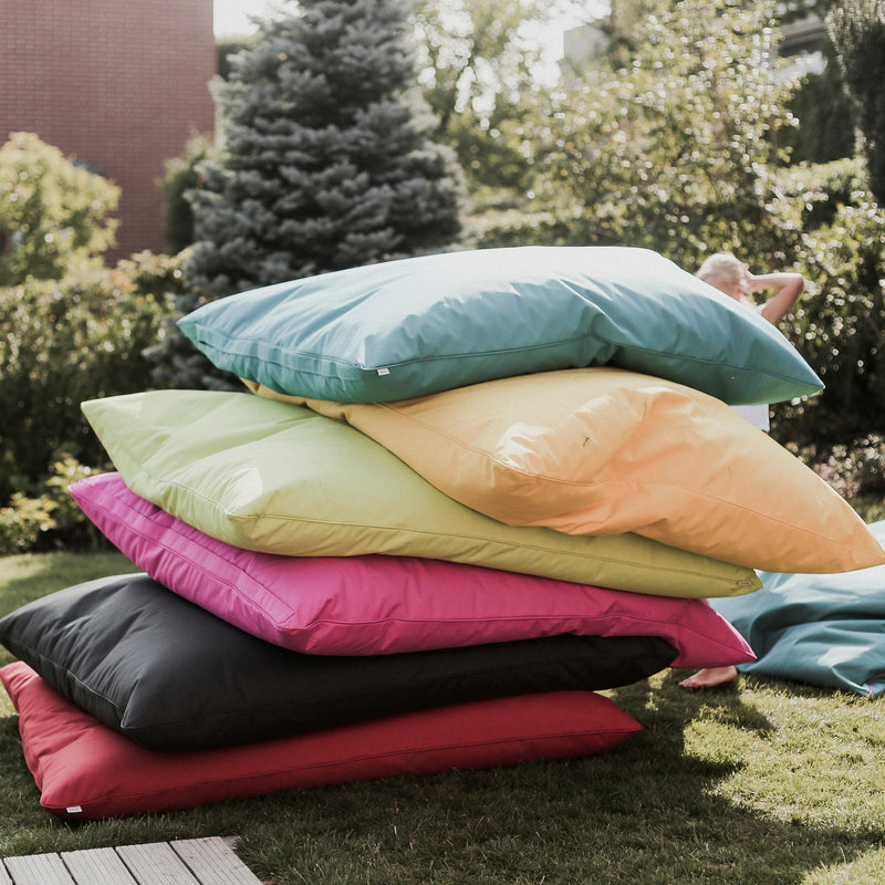 Large, colorful Egoe Vago Bean Bag cushions are stacked in a garden with trees and bushes. These come in shades of green, blue, yellow, pink, red, and black. A person is partially visible behind the stack during an outdoor activity.