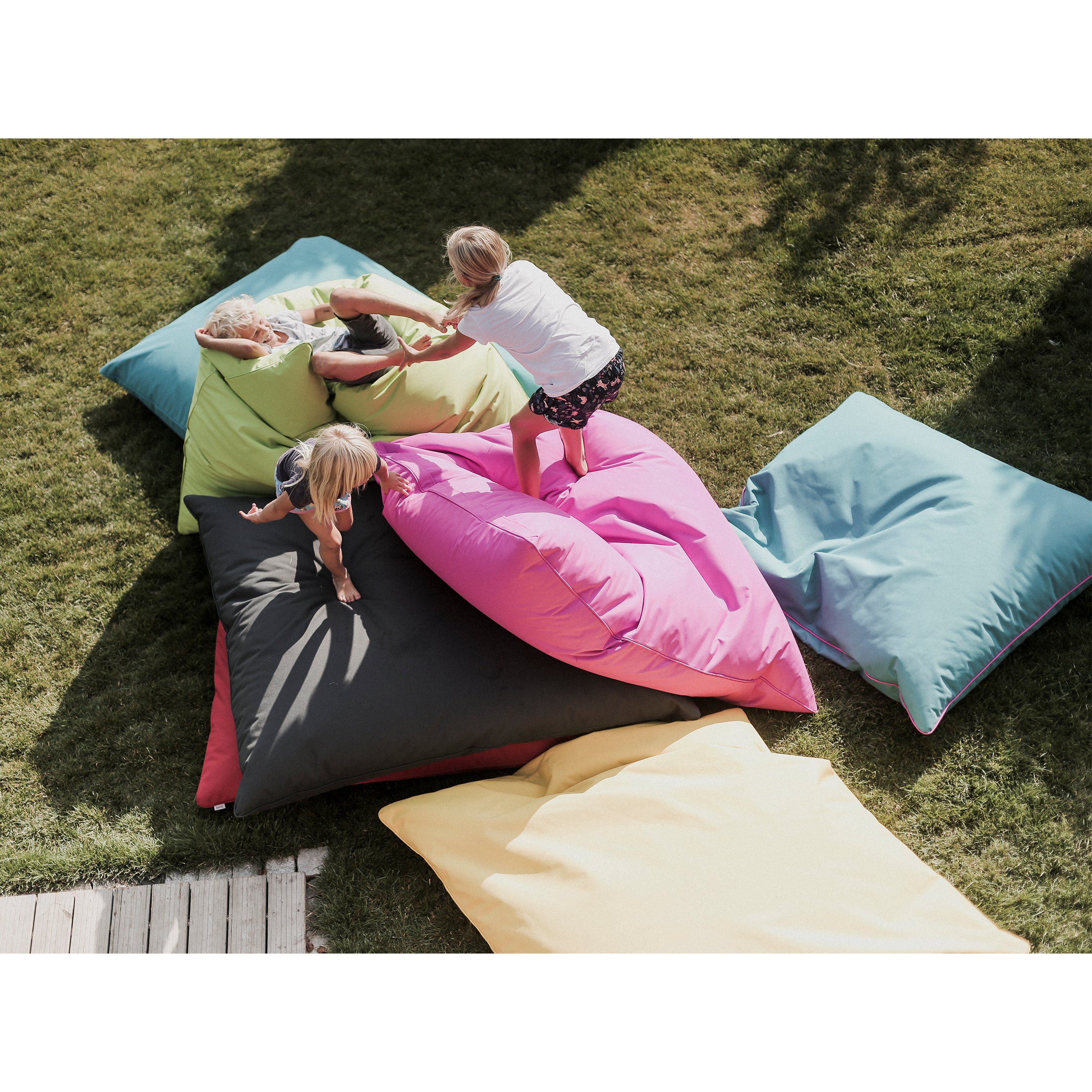 Three children play on a pile of large, colorful Vago Bean Bags by Egoe, made from durable Kortexin fabric. Arranged on grass, the bean bags come in pink, blue, yellow, green, and black. Sunlight casts shadows highlighting this cheerful outdoor activity on a bright day.