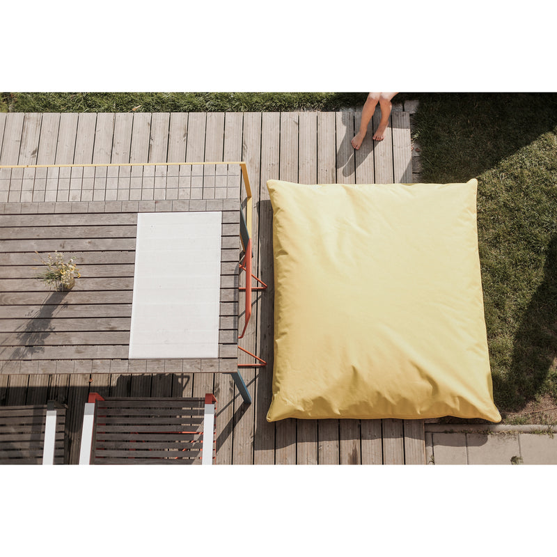 Aerial view of a wooden deck featuring an Egoe Vago Bean Bag in yellow Kortexin fabric next to a rectangular wooden table. Part of a person’s legs appears at the top right corner, suggesting outdoor activity. A small plant in a vase adorns the table, with grass encircling the deck.