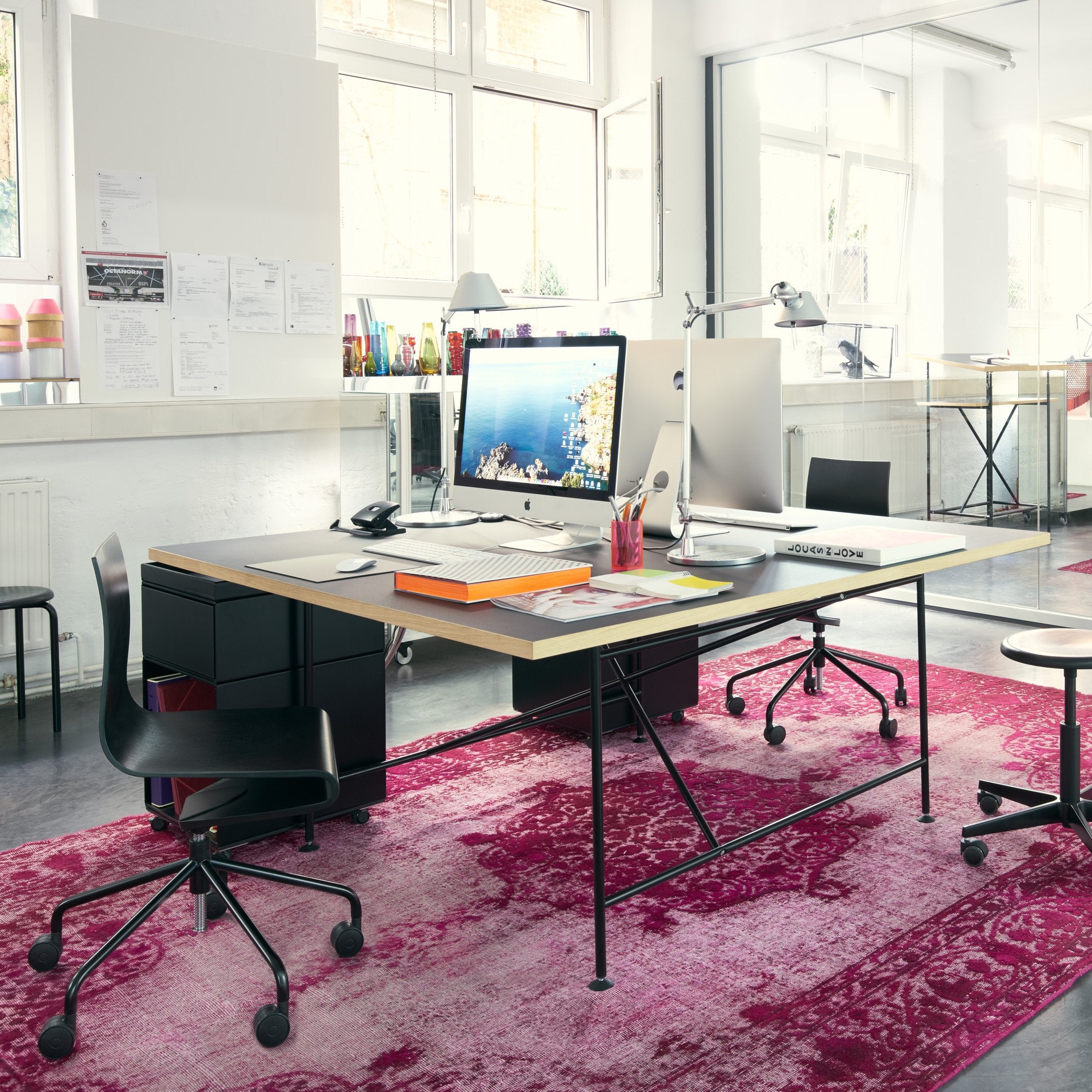 A modern office showcases a sleek Eiermann 1 Conference Table by Richard Lampert, adorned with a computer, notebooks, and pens. Two black chairs surround it on a large pink patterned rug, while bright windows illuminate the design with natural light.