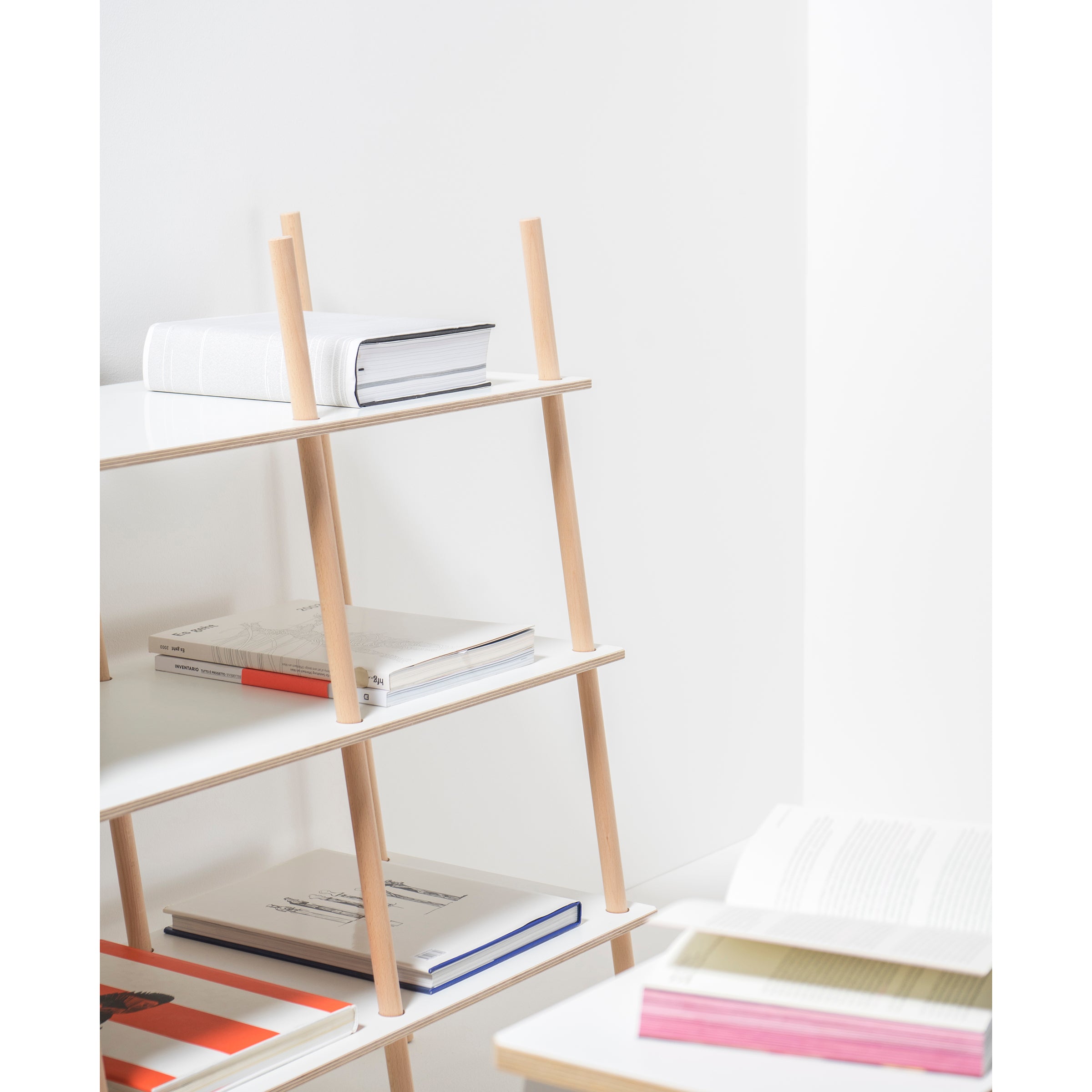 The Es Sideboard by Moormann, crafted from birch plywood with light wooden supports and white shelves, displays a few books in a clean, modern aesthetic. A nearby table holds an open book with colorful pages, enhancing the bright and minimalist scene achieved through precise tolerances.