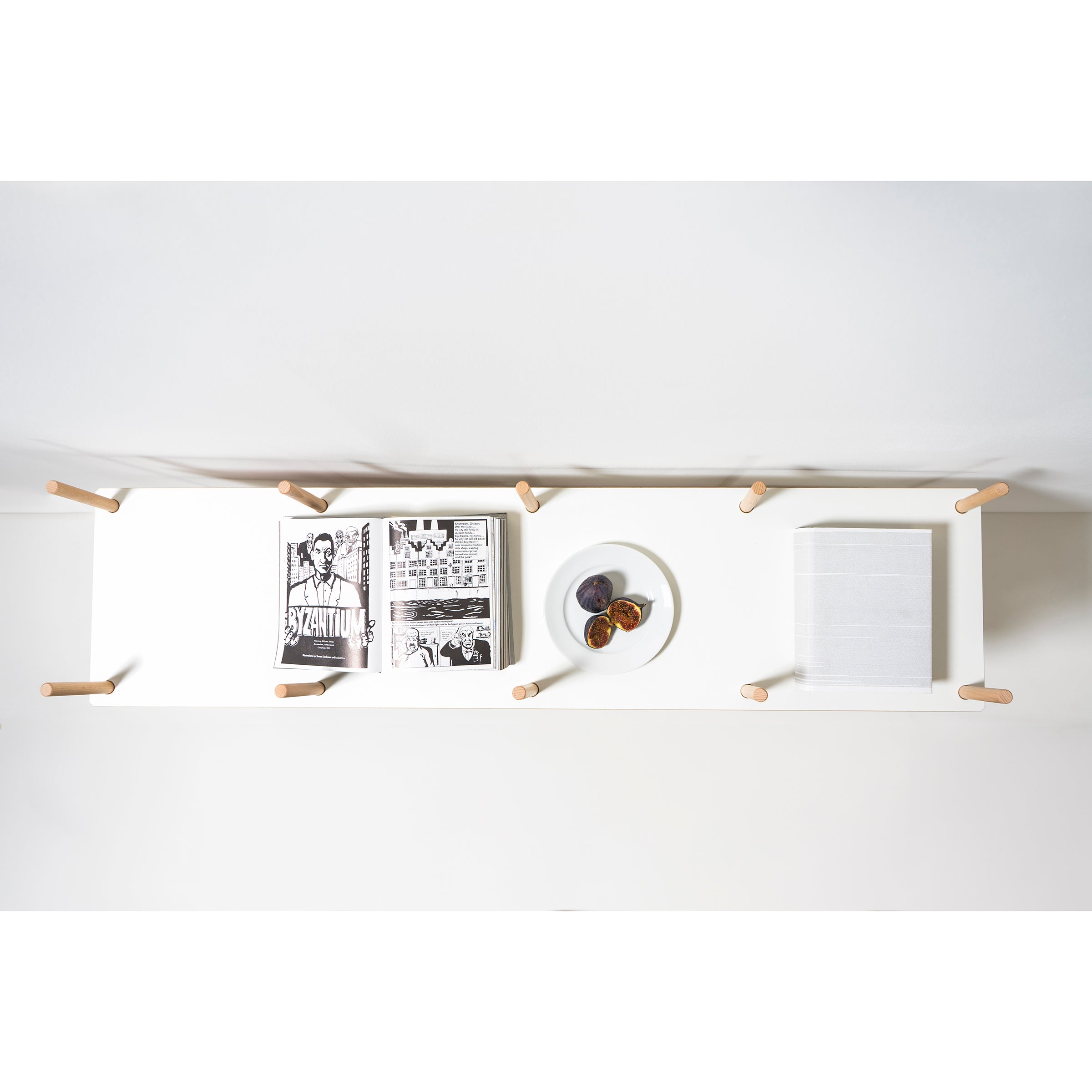 The Es Sideboard by Moormann features a minimalist design with sleek birch plywood legs. Viewed from above, it showcases a stylish arrangement with an open comic book, a closed book, and a round dish for dried fruit or small items, all in an organized layout.