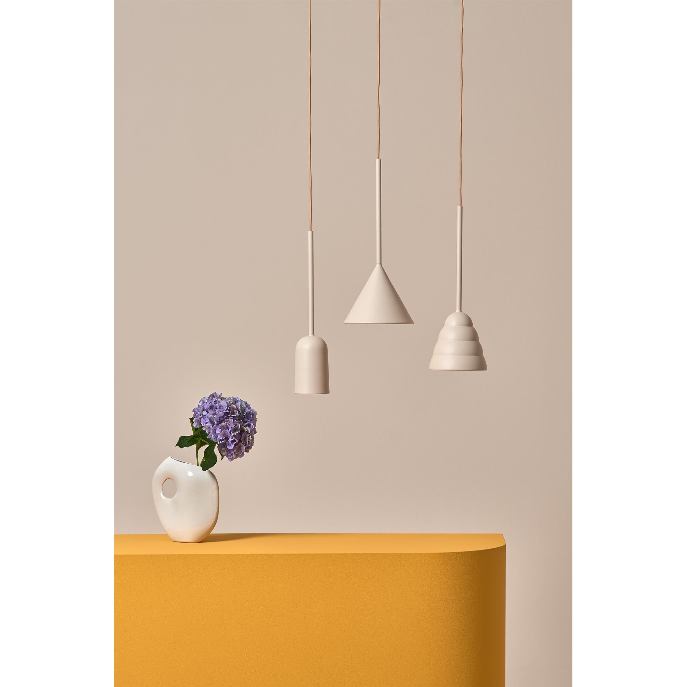 Minimalist design is highlighted with three Schneid Figura Cone Desert Sand pendant lights hanging above an ochre table. A sculptural silhouette forms with a small white vase holding purple hydrangeas against a beige background.