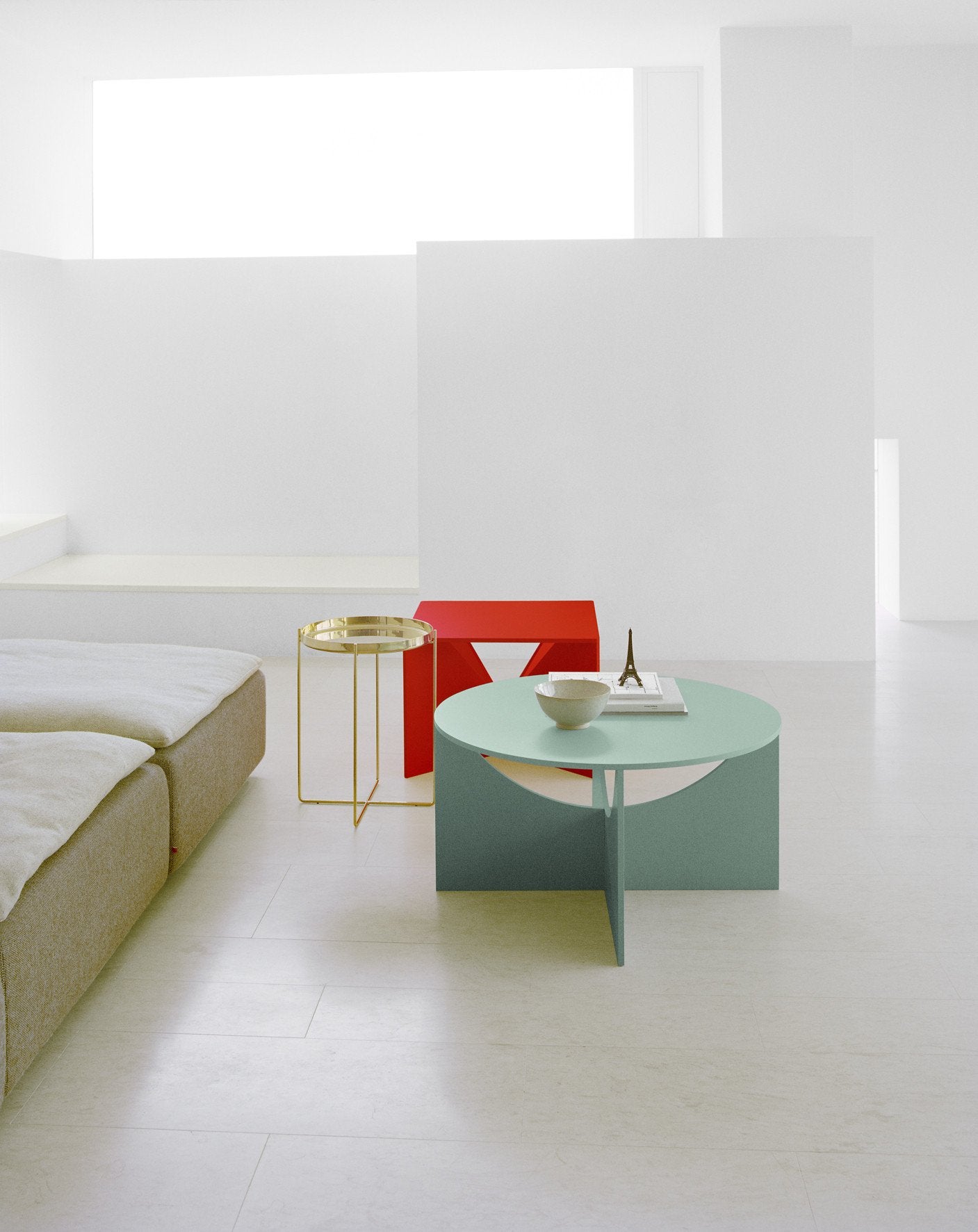 A minimalist living room with pastel tones highlights the FK04 CALVERT pale green coffee table by E15 in a Knock-Down style, surrounded by books, a white bowl, and decor. Nearby are a gold side table and red geometric chair. Natural light brightens the beige sectional sofa and crisp white walls.