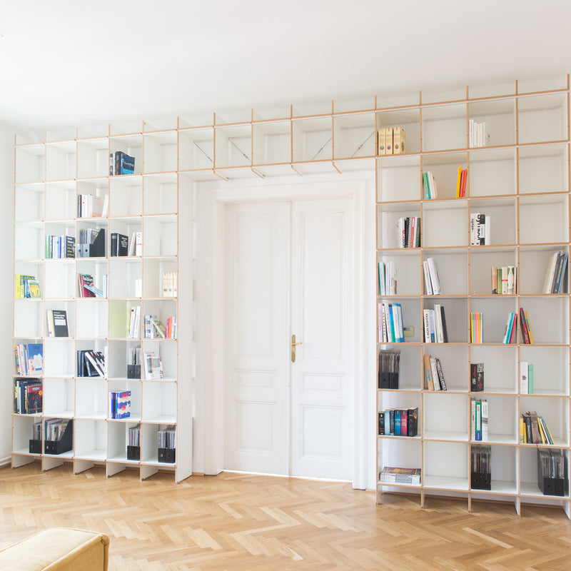 A minimalist living room features a light wooden floor and a white wall with a floor-to-ceiling Moormann FNP shelf framing a white double door. The versatile design ensures the shelves neatly accommodate books and decorative items.
