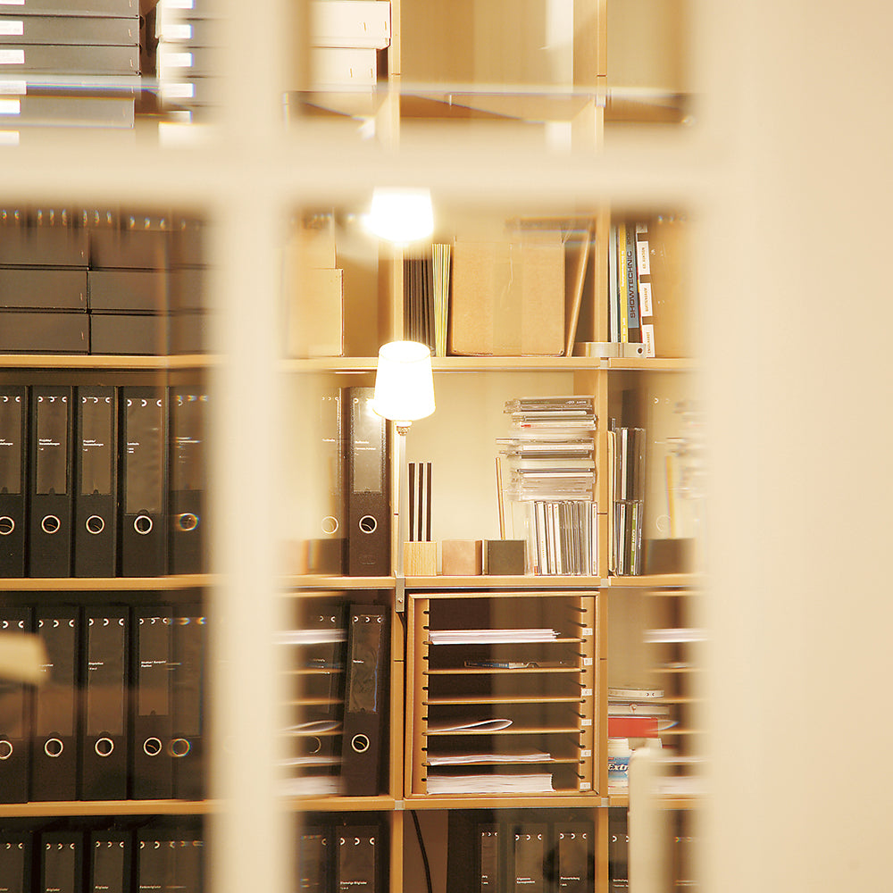 Through the window, a glimpse reveals shelves filled with black binders, boxes, and office supplies. The FNP- and EGAL-shelf system by Moormann ensures order, while the soft glow of the Rosi Lamp adds warmth to the organized workspace.