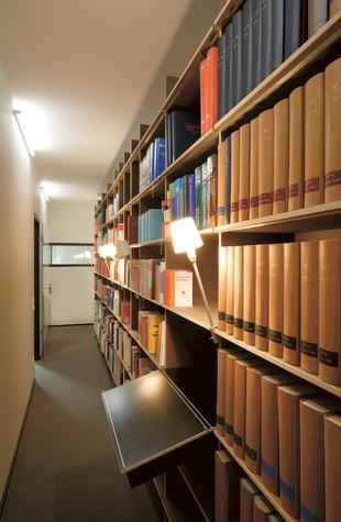 Bookshelves, including the FNP- and EGAL-shelf systems by Moormann, lined with colorful books fill a narrow library corridor. Rosi lamps softly illuminate sections of shelves with a black reading stand attached to one. The hallway leads to a door at the end.