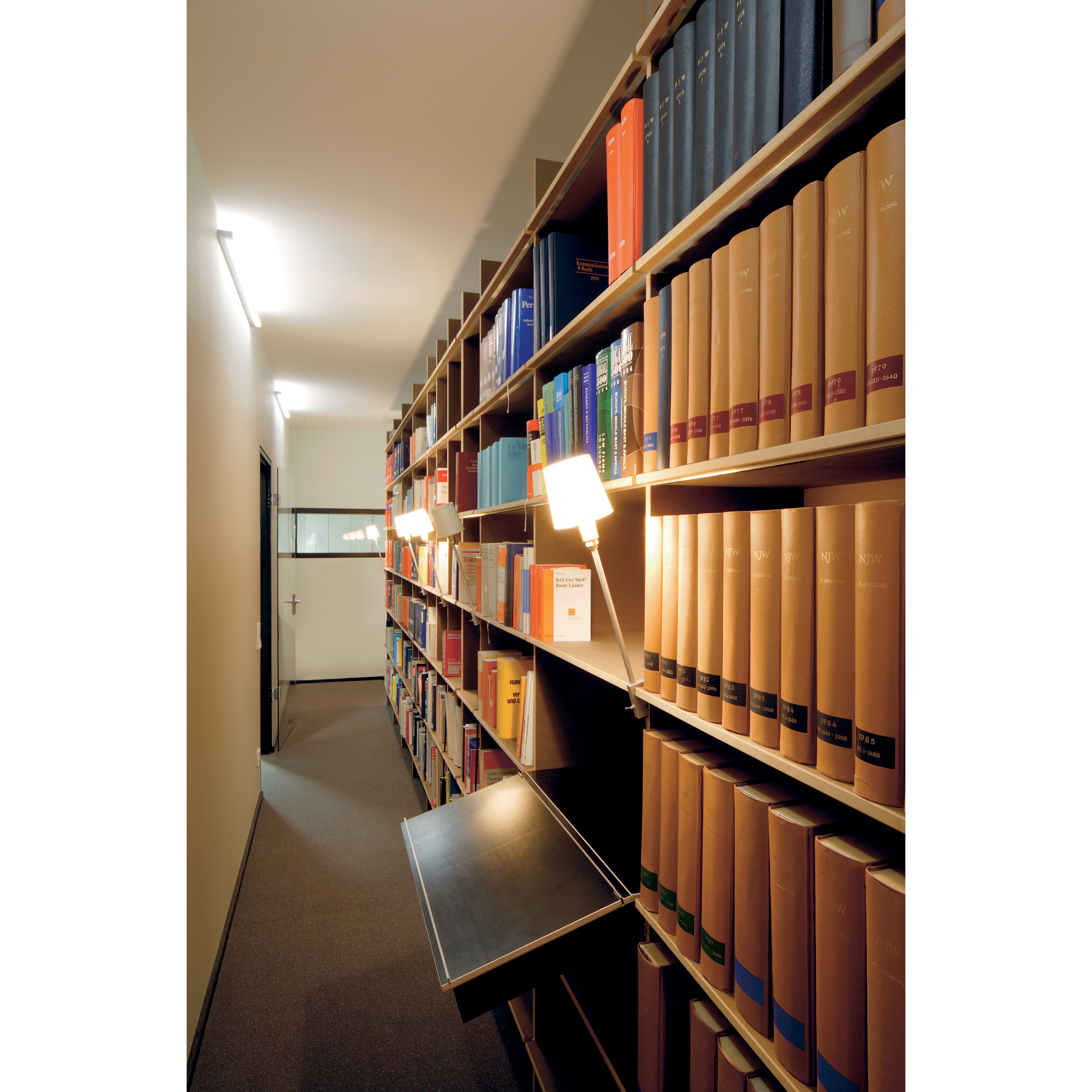 A narrow library aisle features tall FNP and EGAL bookshelves laden with multicolored books. The stylish Rosi Lamps by Moormann illuminate the scene. A window and a closed door are visible at the end of the aisle.