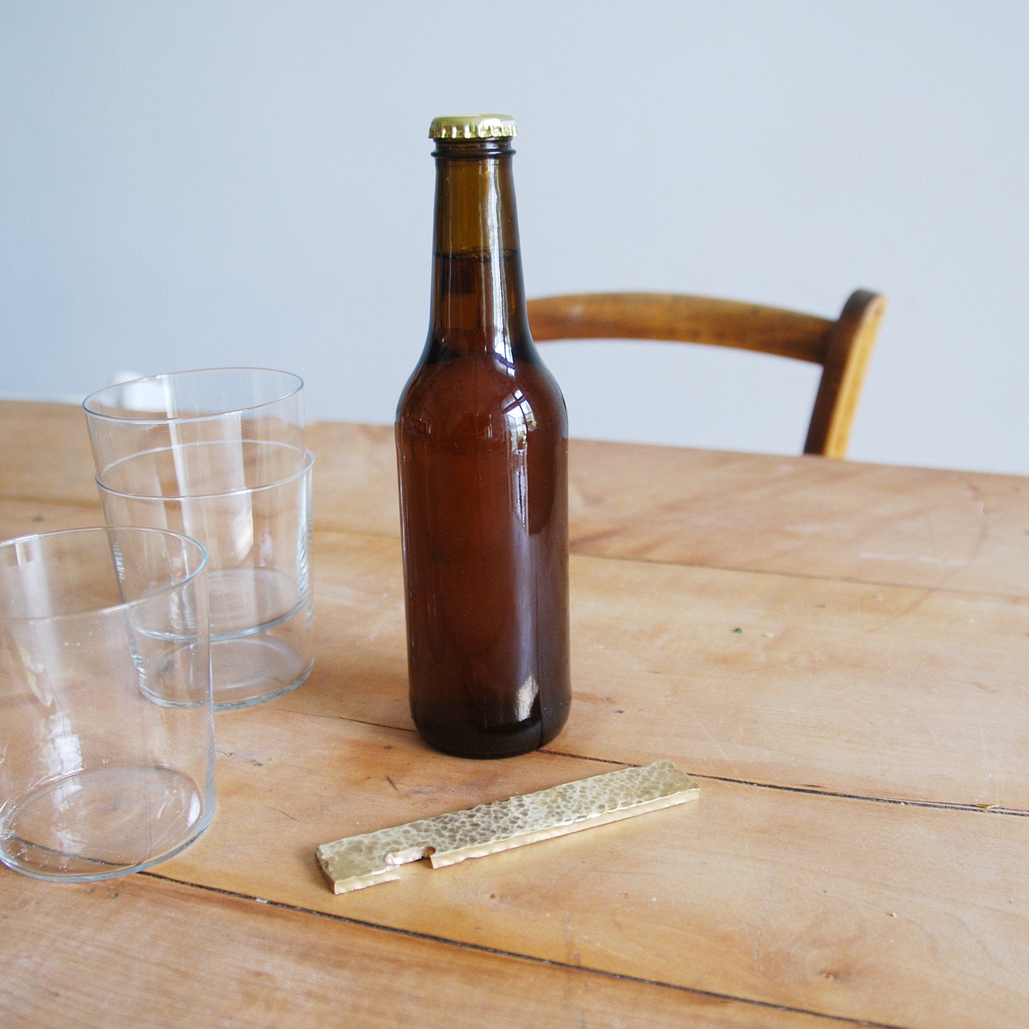 A brown glass bottle with a gold cap is placed on a wooden table beside two empty clear glasses and a brass GoodGoods Open bottle opener. A wooden chair is partially visible in the background.