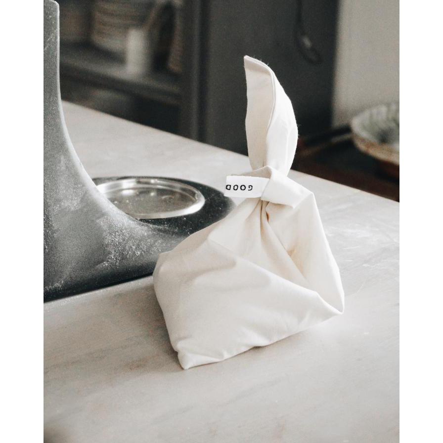 A white Fill it organic cotton bag from GoodGoods, with a tag reading GOOD, is tied at the top and rests on the kitchen counter by the sink. The blurred background of plates and utensils emphasizes its use as a reusable bag for unpackaged shopping.