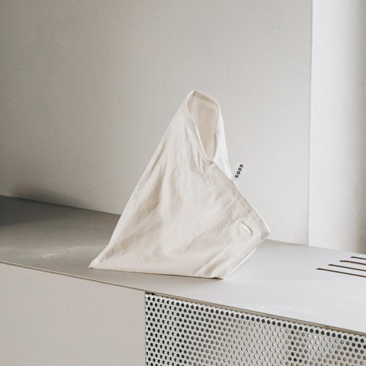 A white fabric object, designed as a minimalist bag made of organic cotton from GoodGoods Fill it collection, lies on a light surface. Its angular shape and small side tag add to its modern aesthetic against a neutral backdrop, ideal for non-packaging shopping.