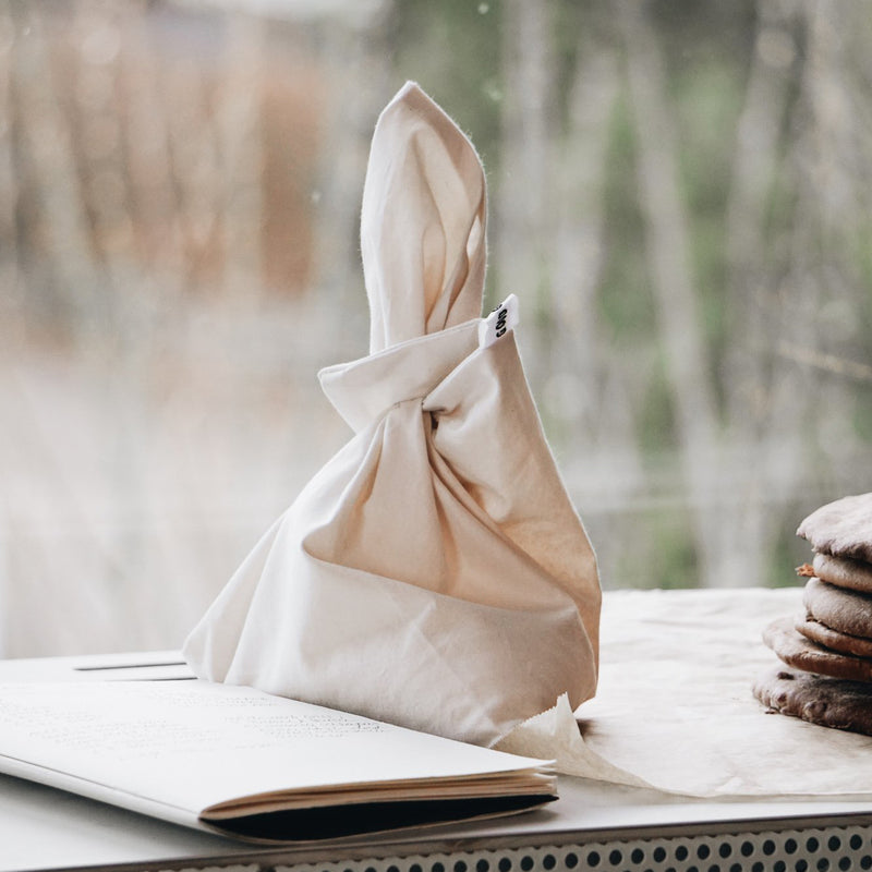 A fabric napkin folded like a rabbit sits next to an open book, alongside organic cotton napkins, against a backdrop of nature through the window—all highlighting the eco-friendly charm of using GoodGoods Fill it reusable bag for non-packaging shopping.