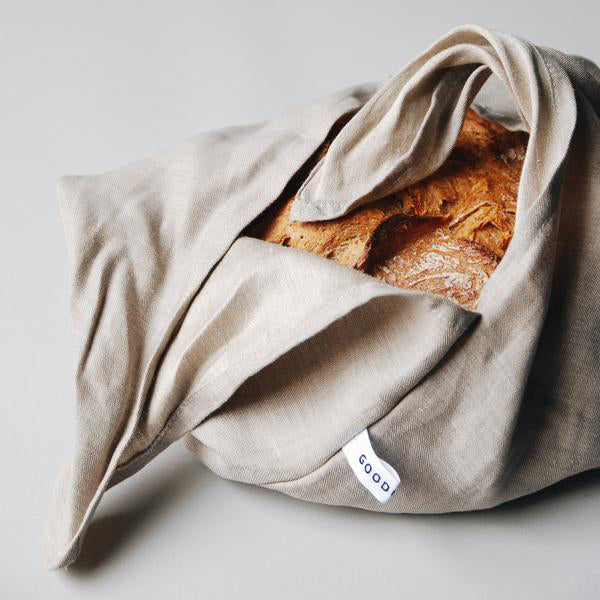 A loaf of crusty bread, partially wrapped in a pure linen cloth, features a small white GoodGoods tag. The golden-brown crust of the Bento bread stands out against the light gray background.