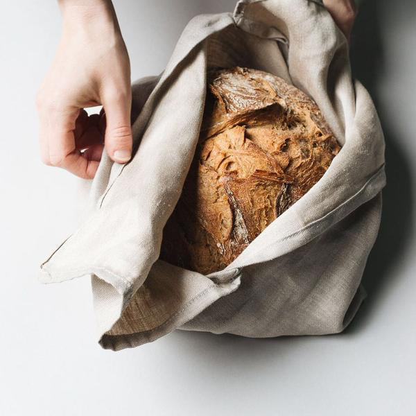 Hands gently unwrap a freshly baked Bento loaf from a pure linen cloth. This rustic bread by GoodGoods has a crusty texture and golden brown color, resting on a light background.