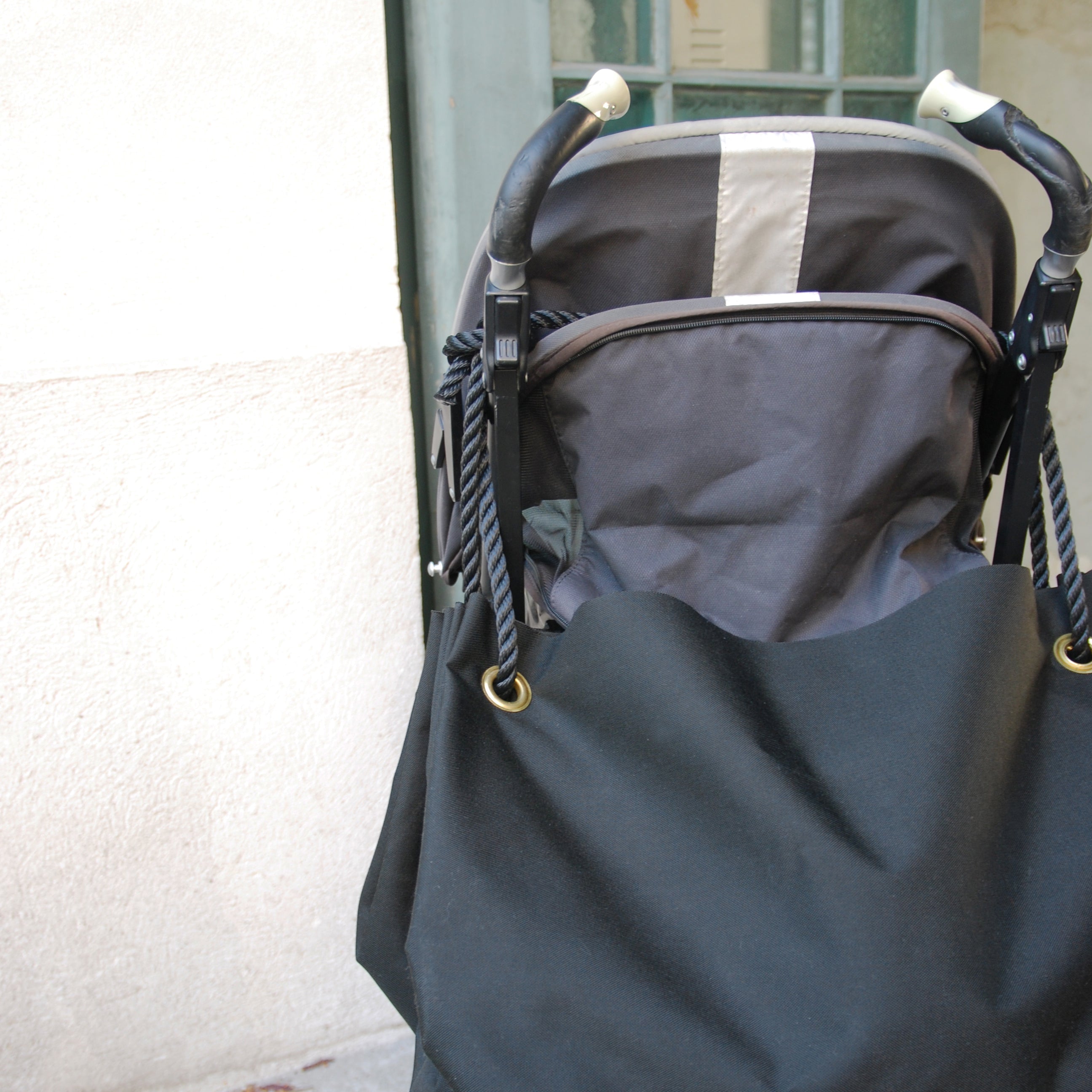 A black stroller with a canopy is parked against a light wall, partially covered by dark fabric. It features black and white handles and holds a large, sturdy GoodGoods Big Bag. A glass window is visible in the background.