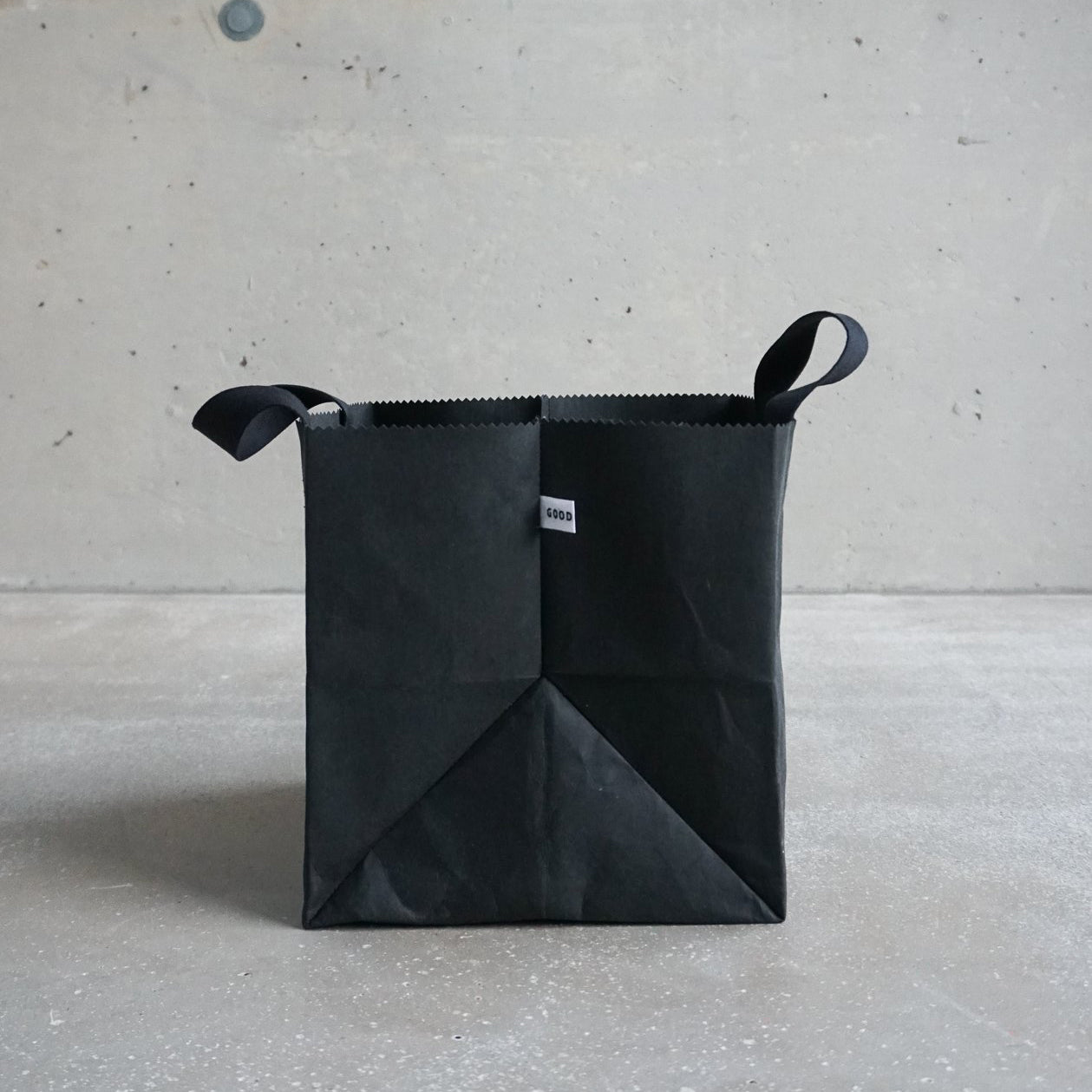 GoodGoods Box Bag, made from foldable vegan leather, has handles and a small white tag on the front. It is placed against a light gray textured concrete wall and rests on a matching light gray floor.
