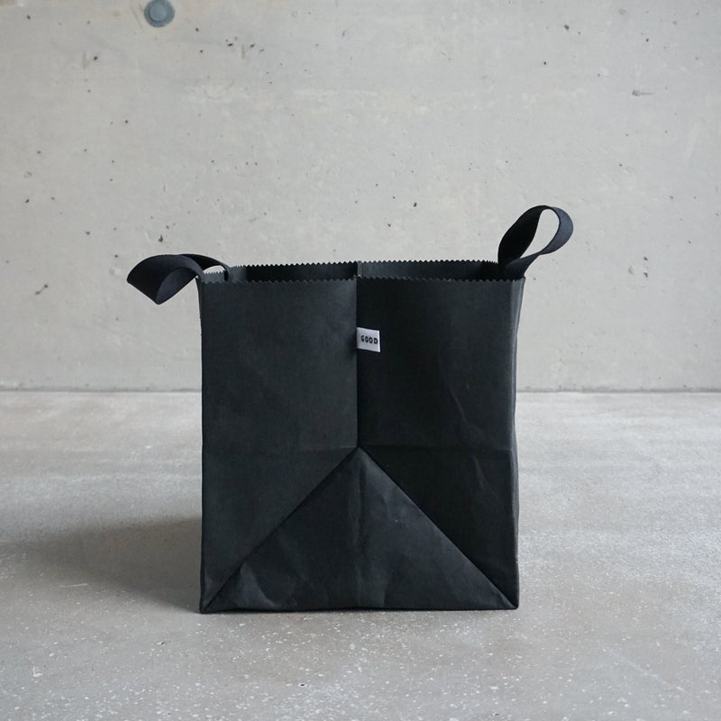 GoodGoods Box Bag, made from foldable vegan leather, has handles and a small white tag on the front. It is placed against a light gray textured concrete wall and rests on a matching light gray floor.