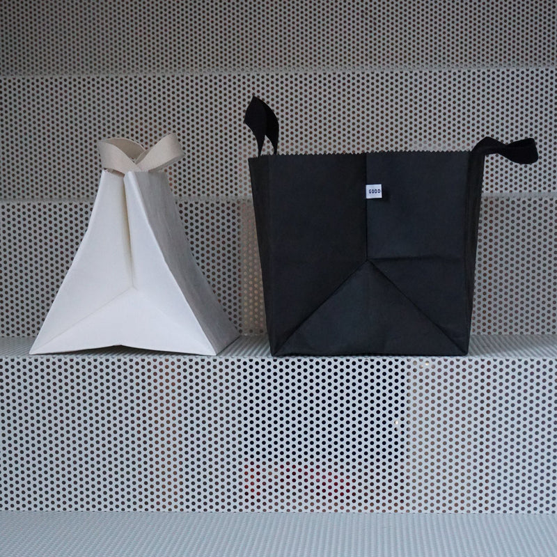 A white triangular tote and a GoodGoods Box Bag in black vegan leather with handles rest on a perforated metal surface.