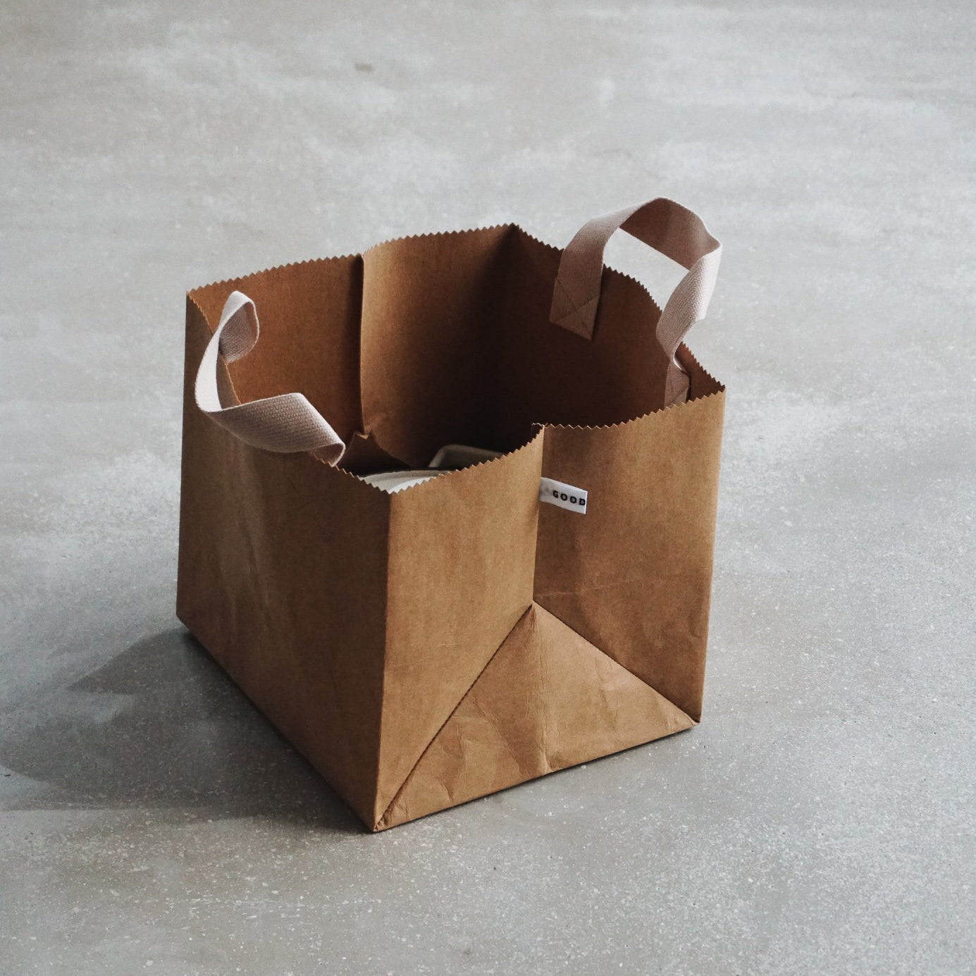 A brown GoodGoods Box Bag, made of vegan leather, featuring two beige handles and a tag on one handle, rests on a gray concrete surface.