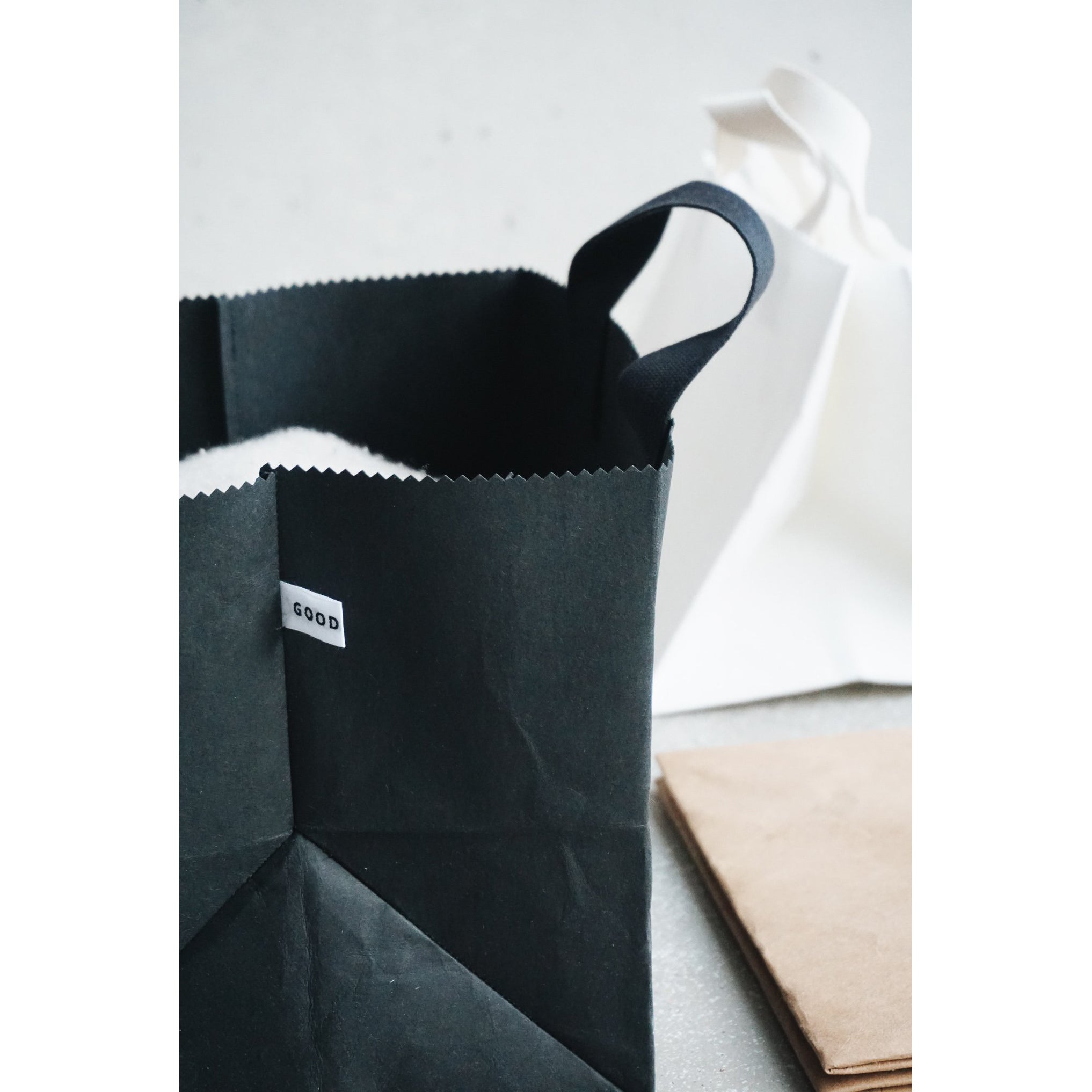 On a light surface, a black Box Bag with handles and a GOOD label by GoodGoods rests beside a white paper bag. Nearby lies folded brown paper, creating a minimalist composition.