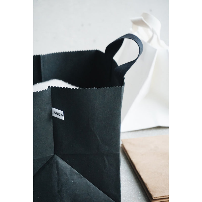 On a light surface, a black Box Bag with handles and a GOOD label by GoodGoods rests beside a white paper bag. Nearby lies folded brown paper, creating a minimalist composition.