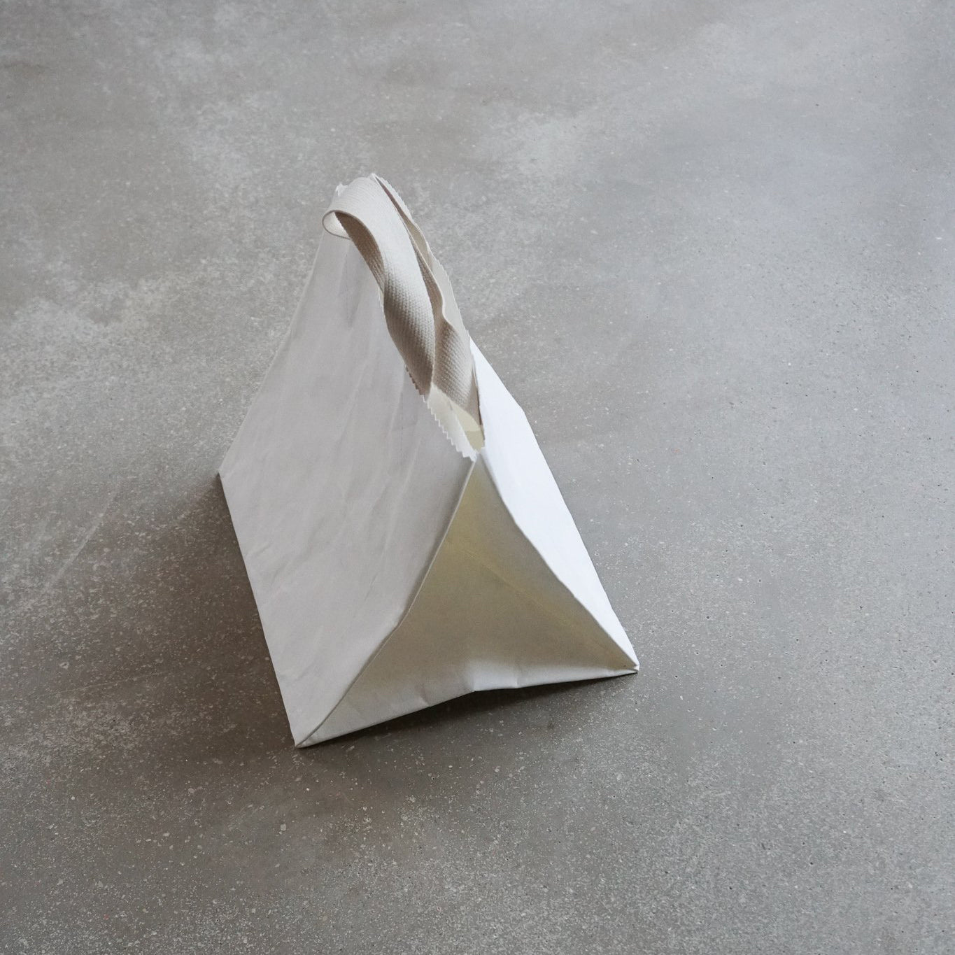 The GoodGoods Box Bag, a white triangular paper bag with a beige handle, resembles a foldable tote on the textured gray surface. Its simple, modern design adds elegance to the scene.
