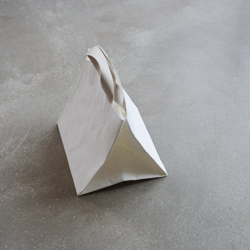 The GoodGoods Box Bag, a white triangular paper bag with a beige handle, resembles a foldable tote on the textured gray surface. Its simple, modern design adds elegance to the scene.