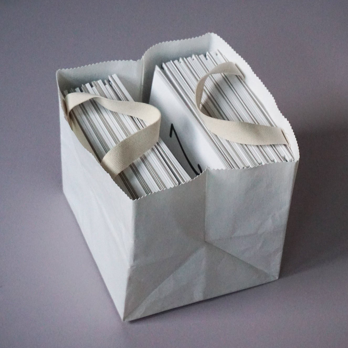 The GoodGoods Box Bag, a chic tote made from foldable vegan leather, features contrasting light handles and is elegantly filled with envelopes on its smooth grey surface.