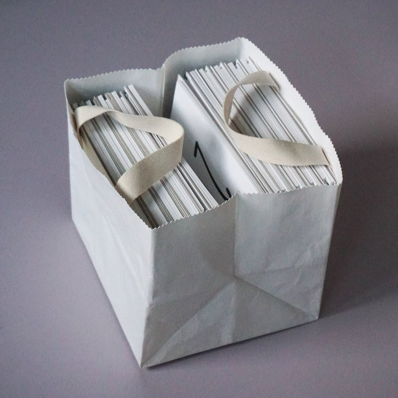 The GoodGoods Box Bag, a chic tote made from foldable vegan leather, features contrasting light handles and is elegantly filled with envelopes on its smooth grey surface.