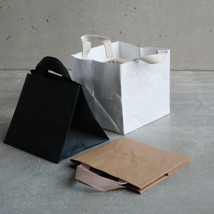 Three bags from GoodGoods are on a concrete floor: a black triangular paper bag, a white square tote with handles, and a folded brown vegan leather bag. The backdrop is a smooth gray wall.