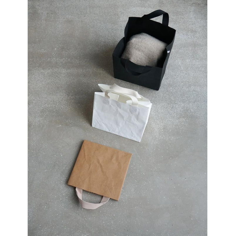 A Box Bag by GoodGoods, made of vegan leather, holds a soft gray item on a gray surface. Below it is a white foldable tote bag resting on a flat brown paper bag.
