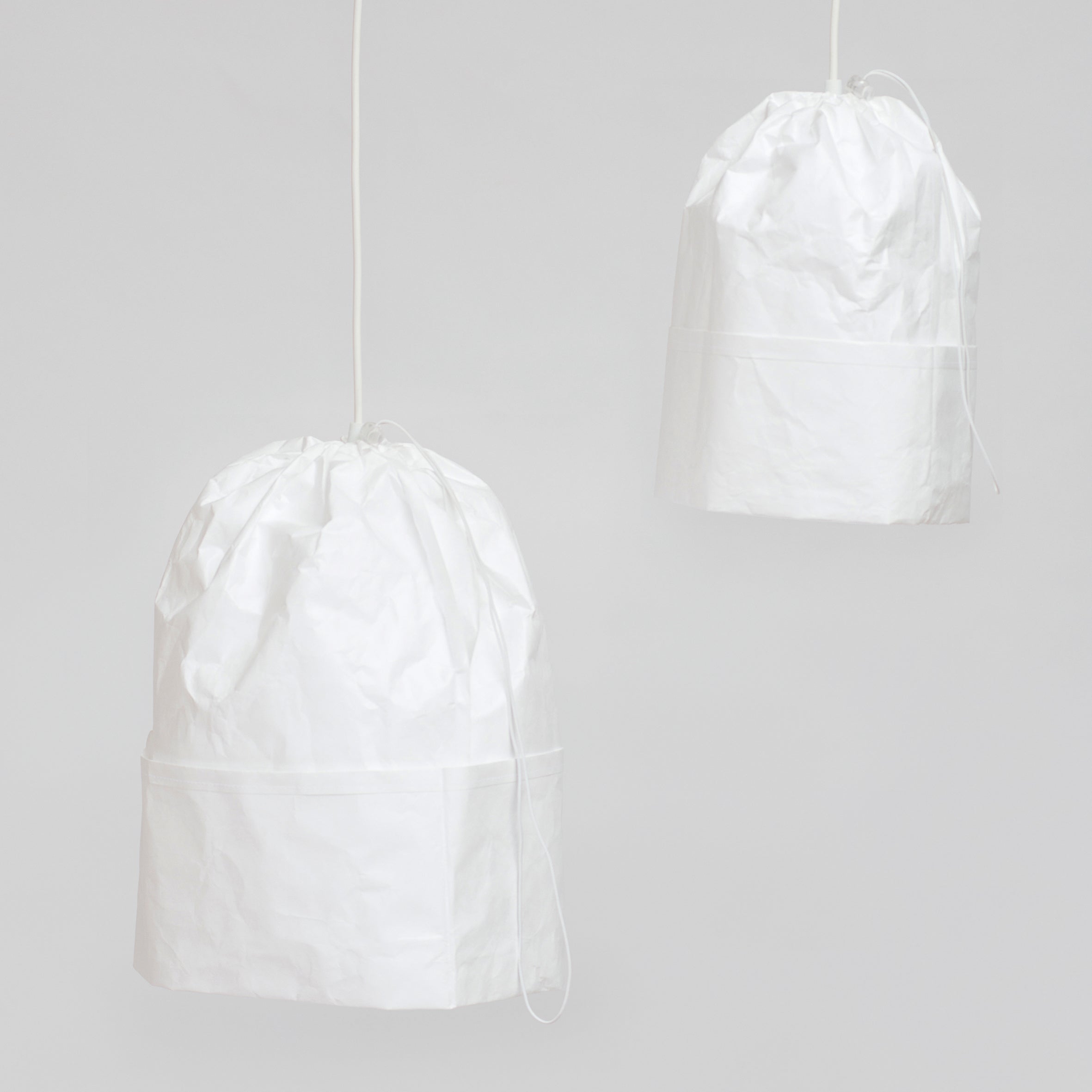 Two Dress a Bulb pendant lamps by GoodGoods hang from the ceiling with Tyvek lampshades resembling crumpled paper bags. Featuring a minimalist design, the soft, matte texture with a drawstring at the top is set against a plain, light gray background.