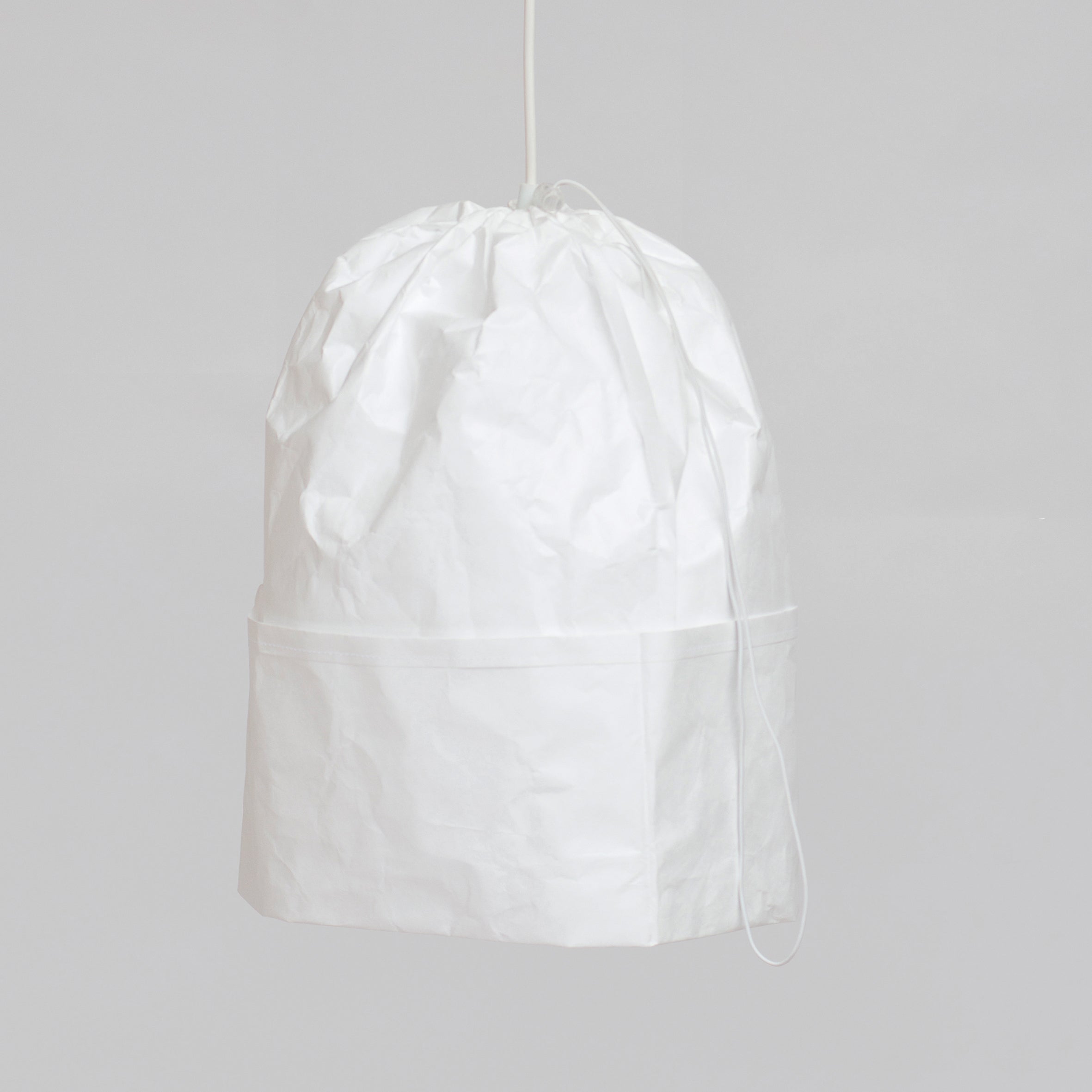 The Dress a Bulb by GoodGoods, a crumpled white Tyvek lampshade with a drawstring design, elegantly hangs against a light gray background, showcasing innovative materiality.