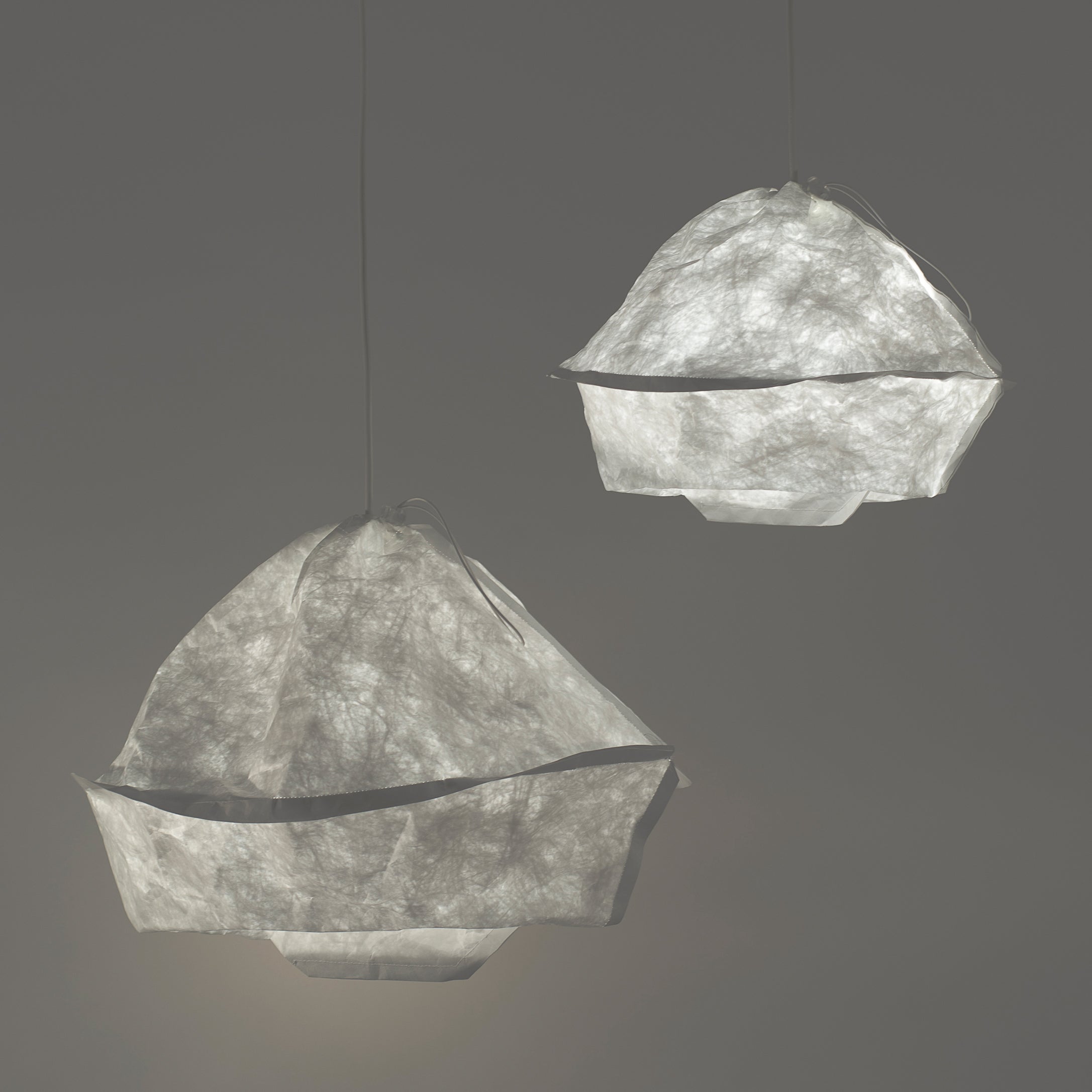 Two Dress a Bulb pendants from GoodGoods, featuring textured white Tyvek lampshades, hang against a gray backdrop. Their organic shapes mimic delicate paper or fabric, highlighting innovative materiality and providing a gentle, diffused glow.