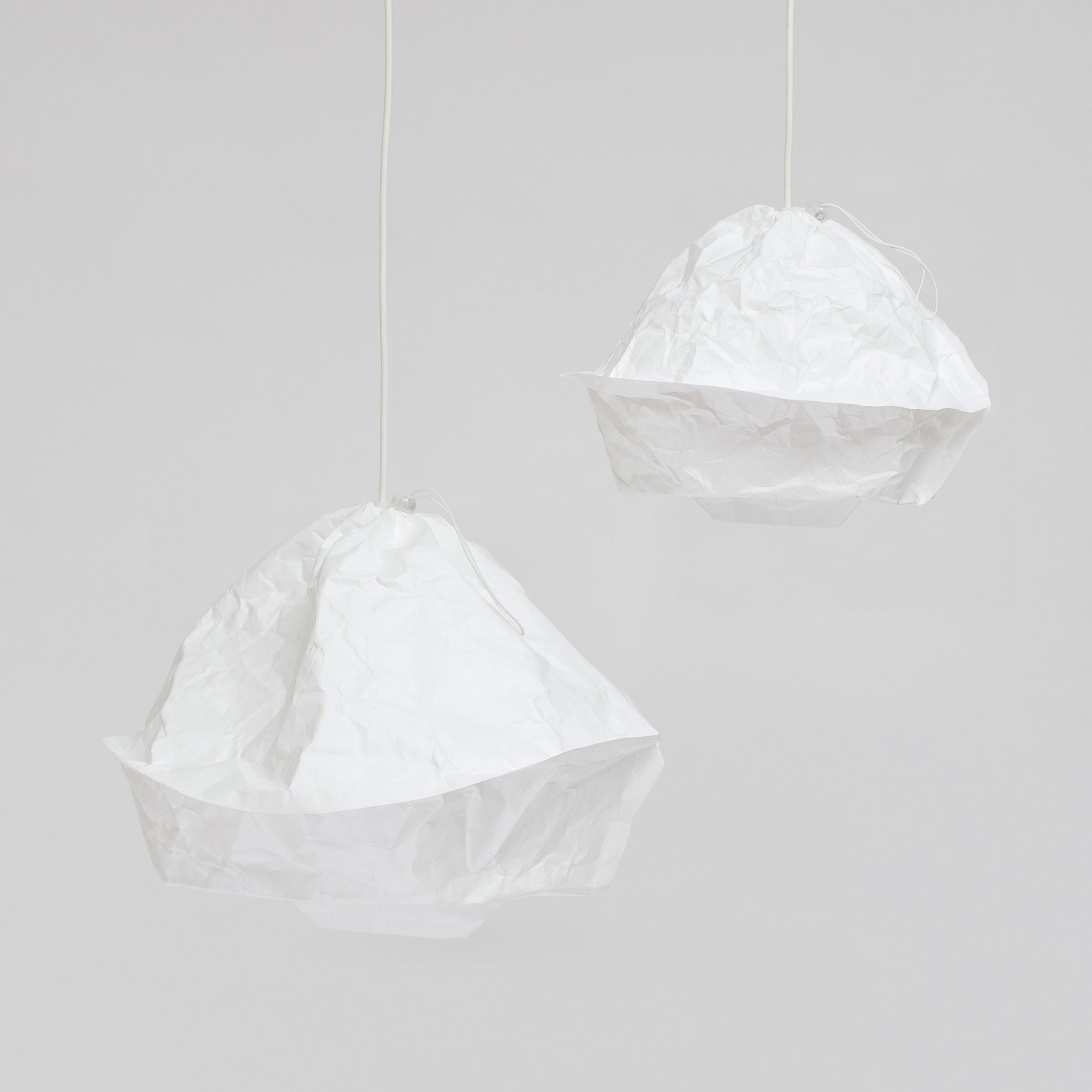 Two Dress a Bulb pendant lamps by GoodGoods, featuring crumpled white Tyvek shades against a gray background. Their irregular shapes lend a playful, artistic look that highlights their innovative material.