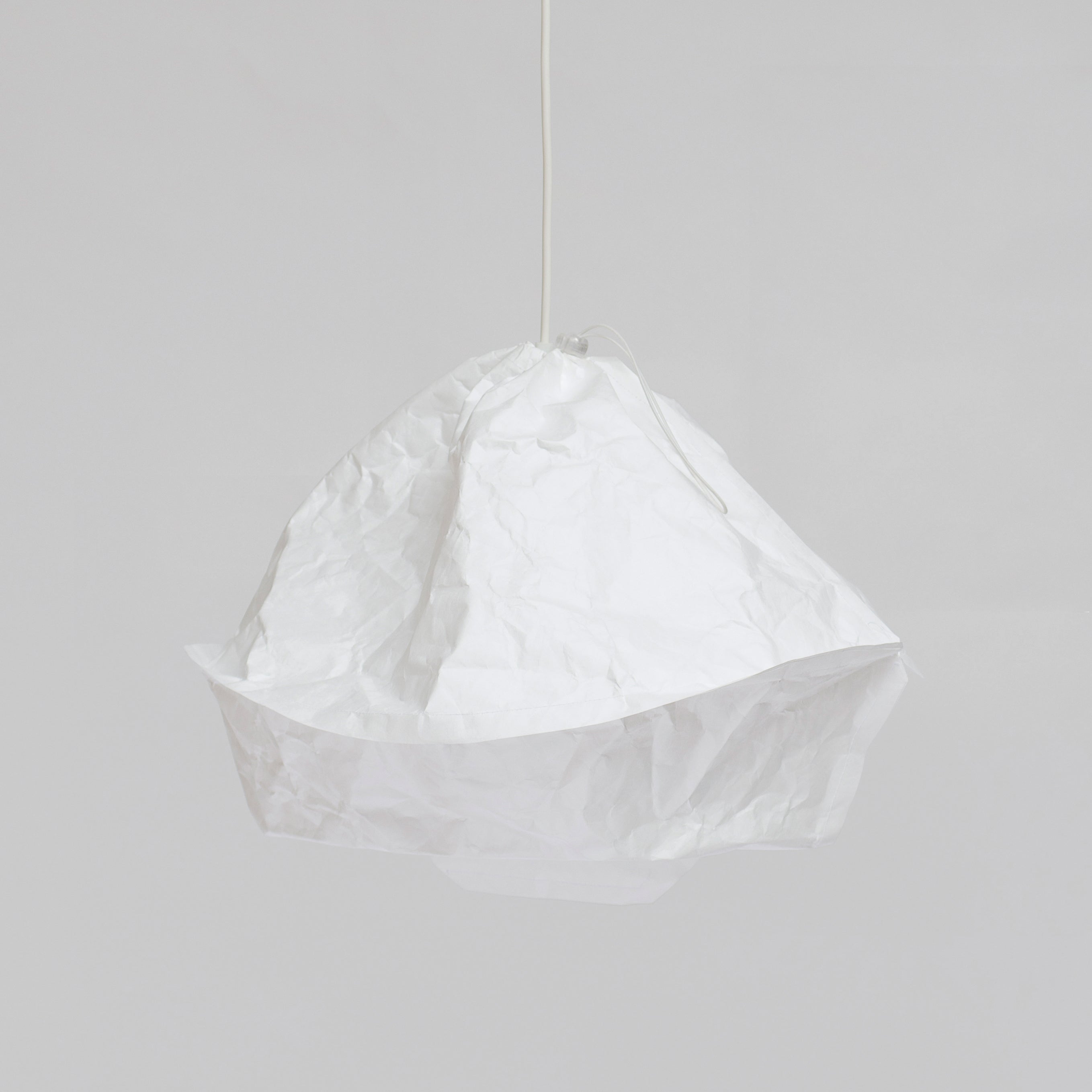 The Dress a Bulb by GoodGoods is a white pendant light with a texture like crumpled paper, set against a gray background. Made from innovative materiality, it resembles a loosely gathered sack, offering a modern aesthetic similar to Tyvek lampshades.