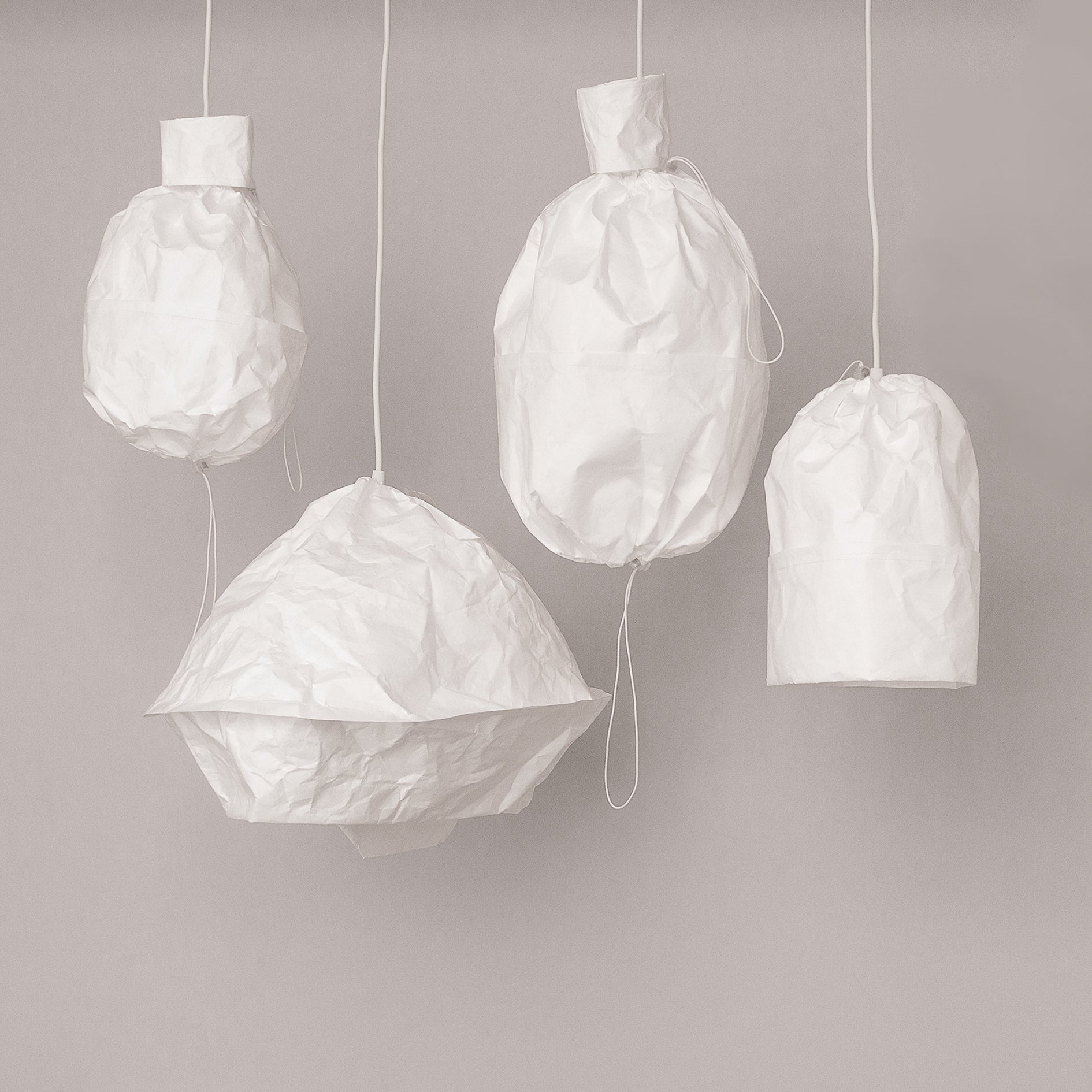 Four Dress a Bulb lanterns from GoodGoods, crafted from crumpled white Tyvek in various shapes and sizes, hang artistically against a plain background. Suspended by thin cords, these lampshades create a minimalist display that showcases innovative materiality.
