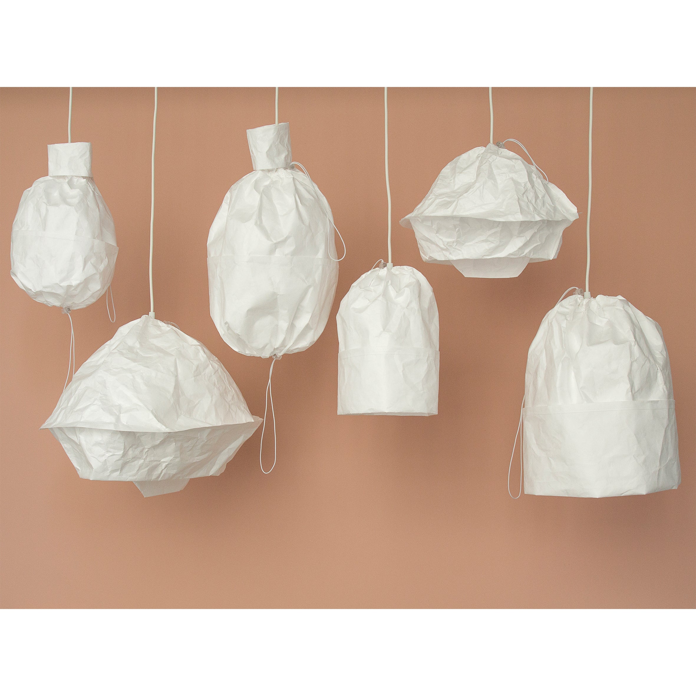 Six crumpled white Dress a Bulb lanterns by GoodGoods hang from the ceiling against a soft beige background. Each flat-packed Tyvek shade is suspended by a thin white cord, creating an artistic and minimalistic display of varying shapes.