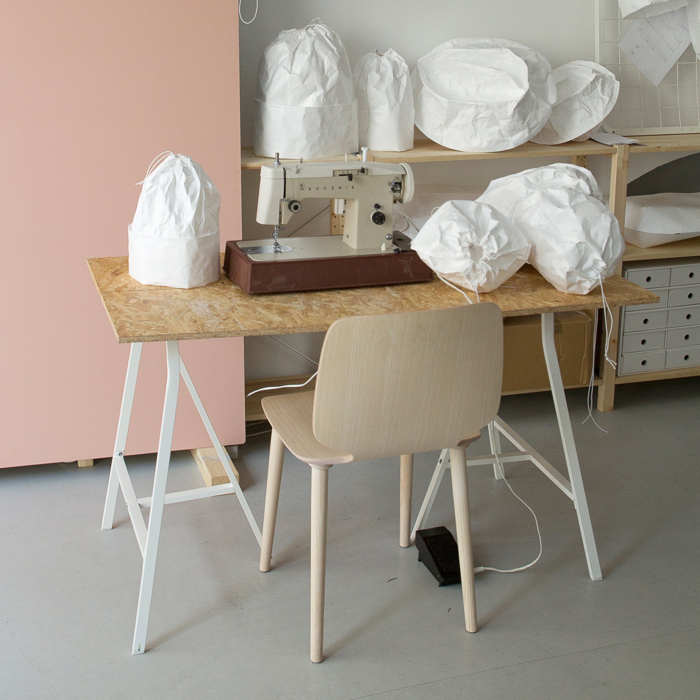 In a cluttered workspace, GoodGoods’ Dress a Bulb Tyvek lampshades sit alongside white fabric shapes on a wooden table with a sewing machine. A light wooden chair is in front of the table, while shelves holding more fabric pieces showcase innovative materiality.